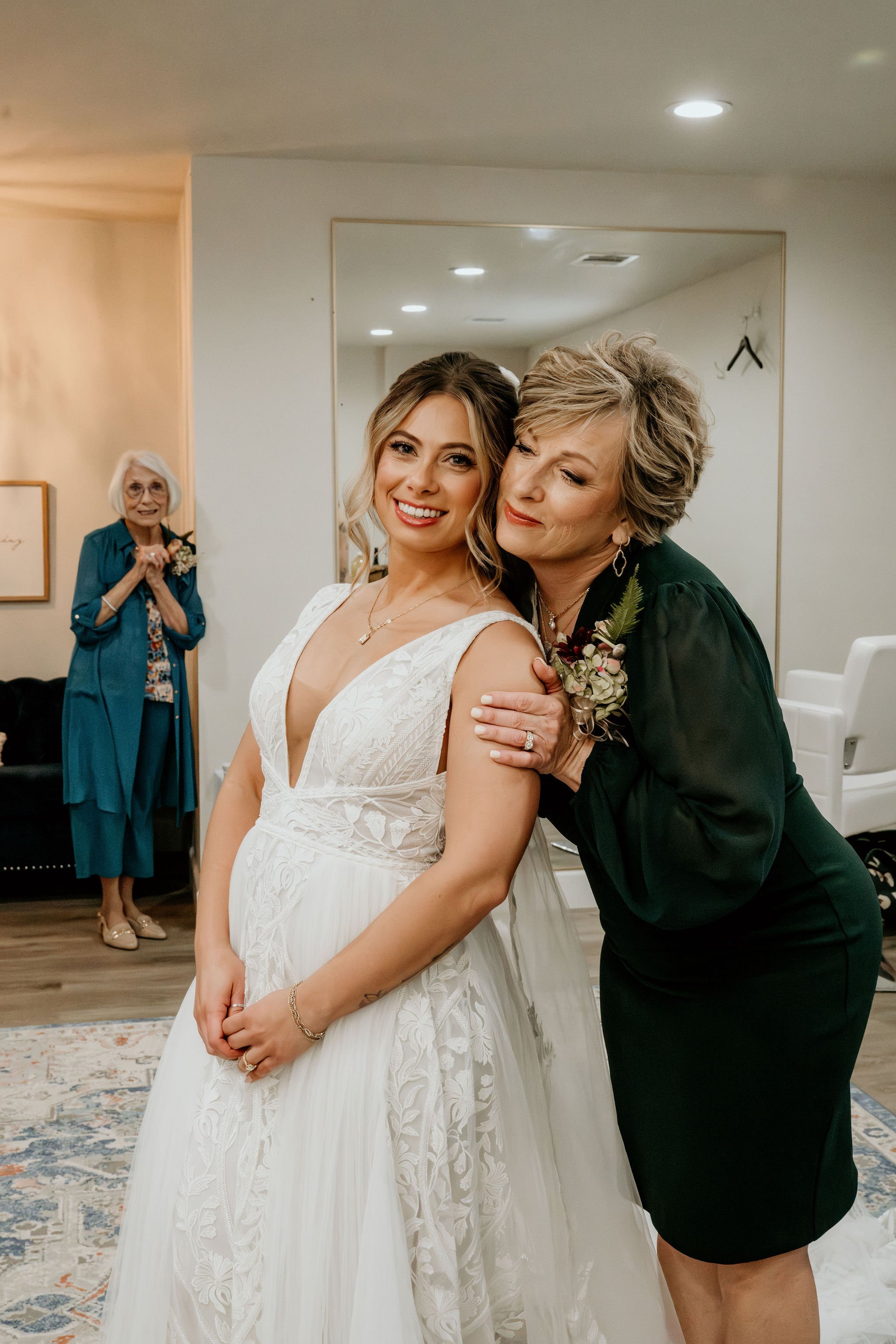 A woman in a white wedding dress smiling as she gets a hug from an older woman in black attire, with another older woman in teal in the background indoors.