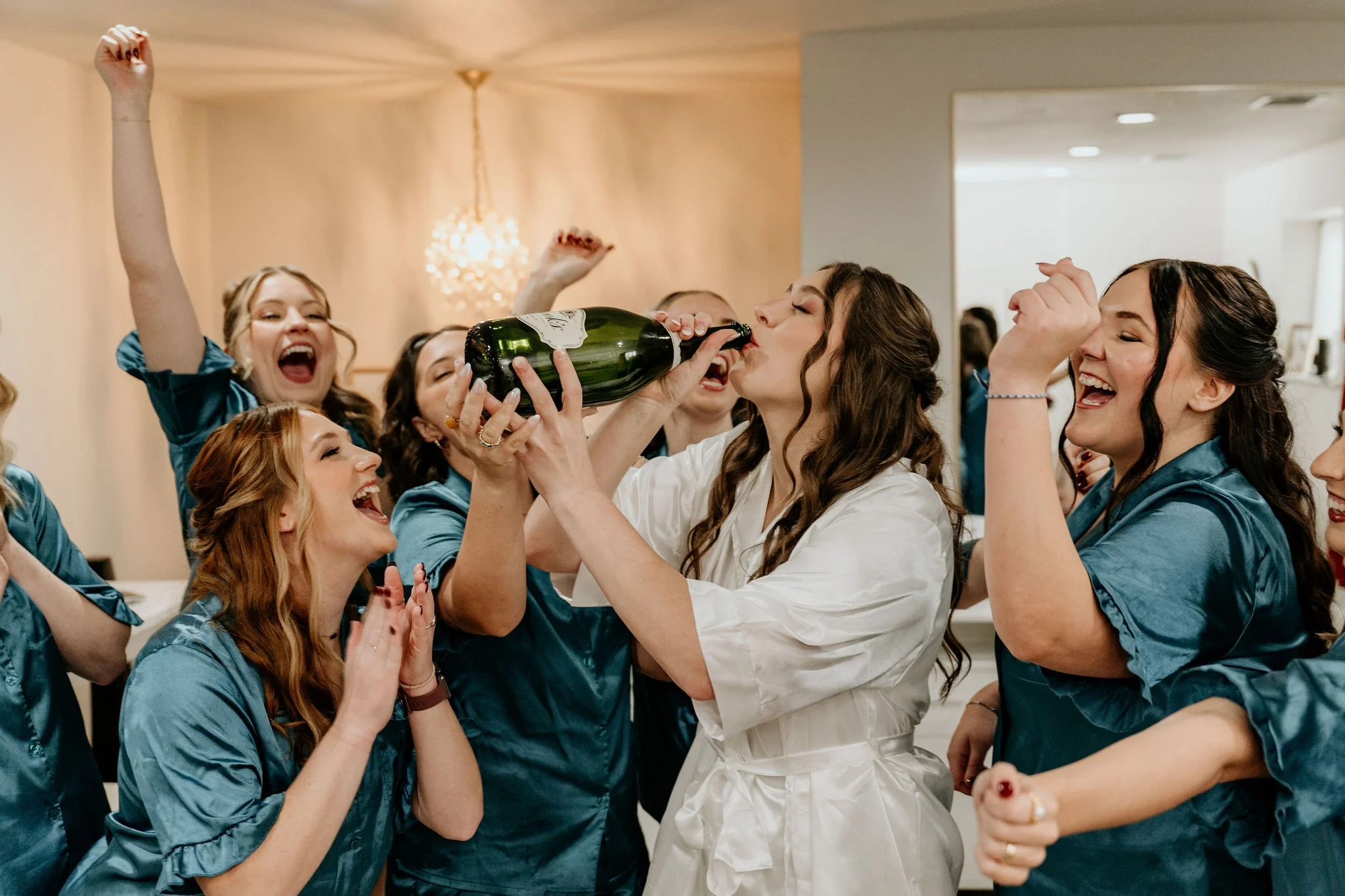 A group of women celebrating and drinking champagne at an event, with some women in matching blue robes and one in white, in a decorated indoor setting.