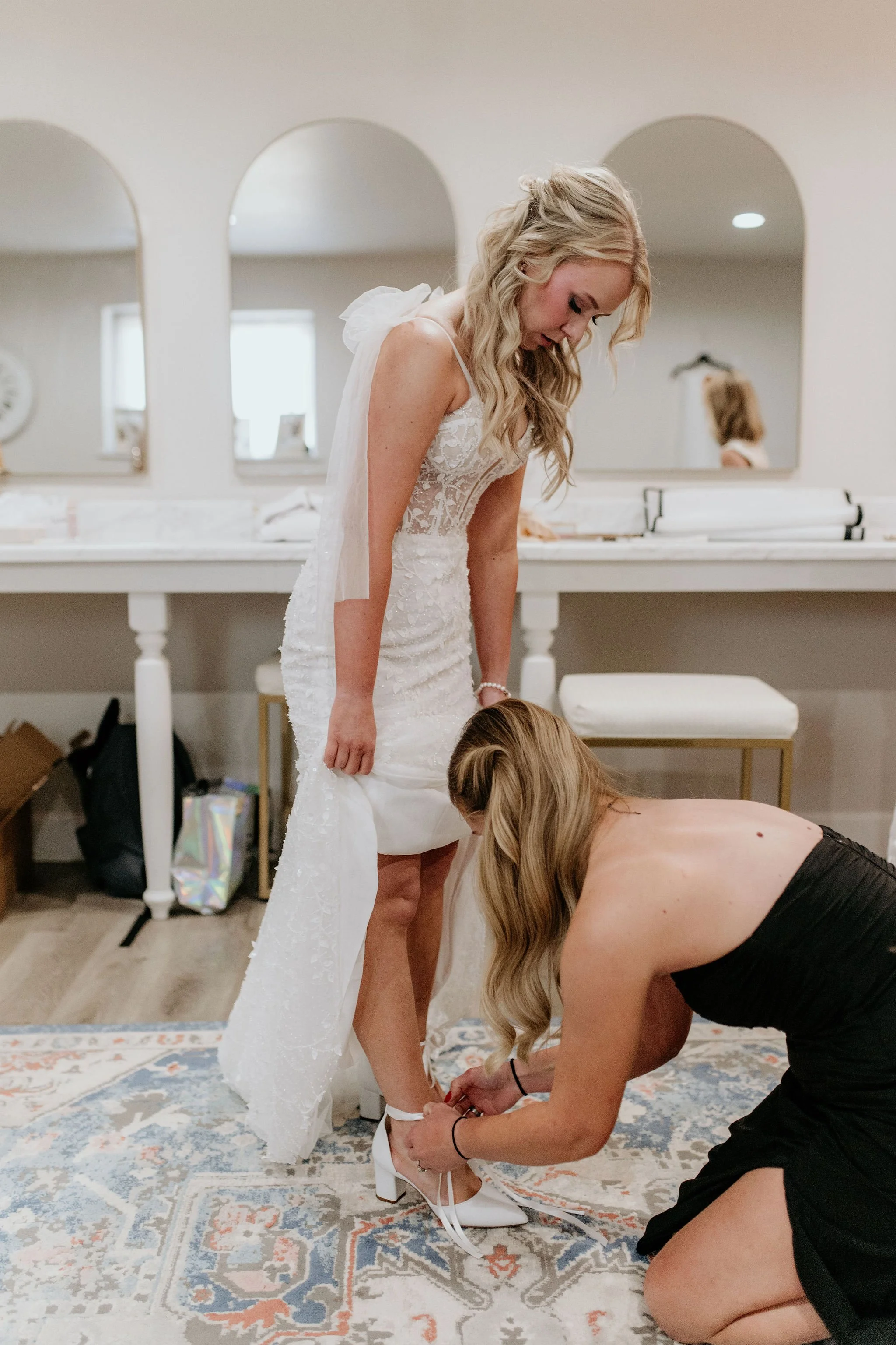 A bride in a lace wedding dress stands while another woman helps her put on white high heels in a room with a large mirror and a decorative rug.