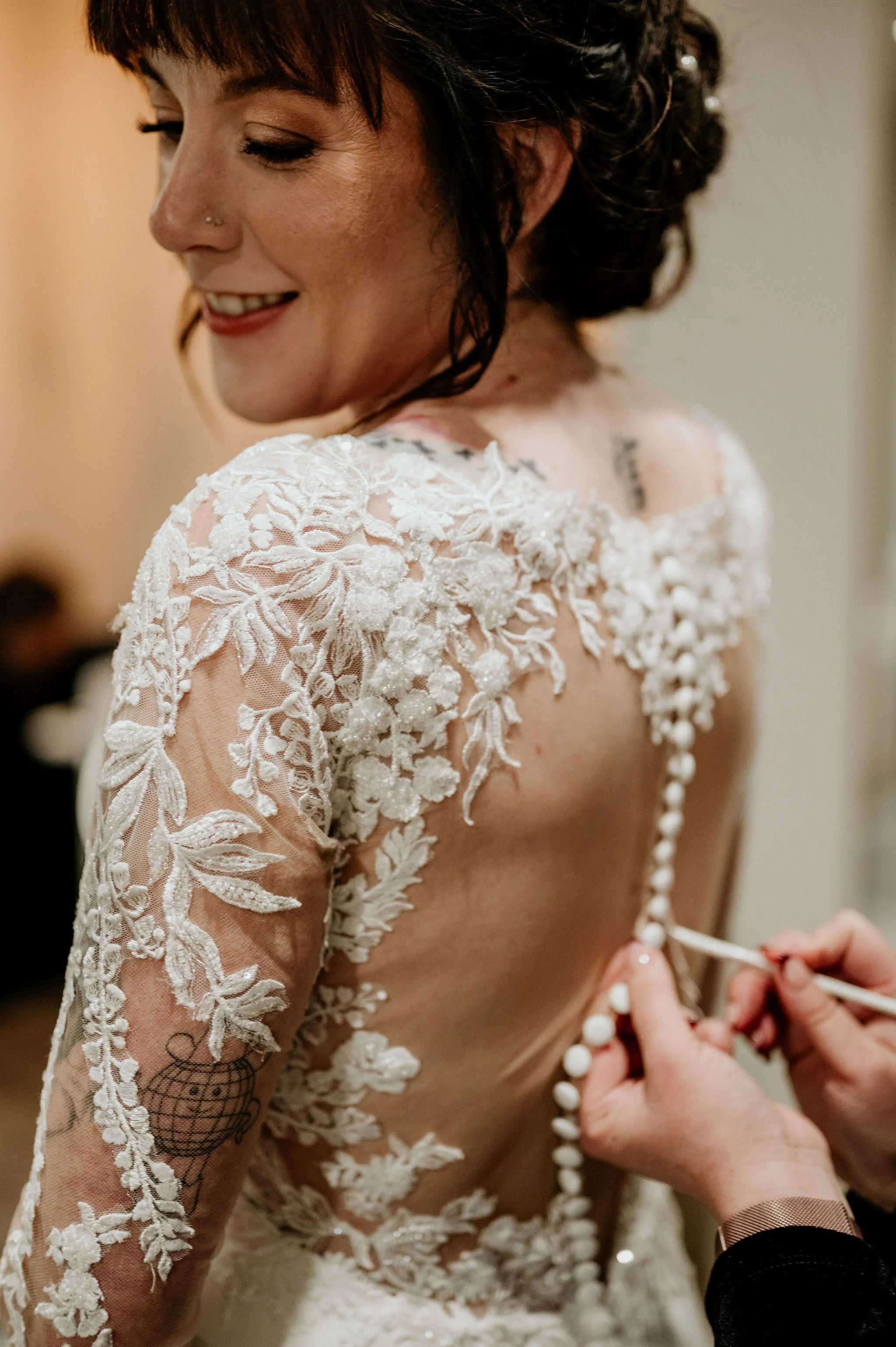 A bride with dark hair and a nose piercing is getting her wedding dress buttoned up. The dress is white with lace and floral embroidery on the back and sleeves. She is smiling and looking down.