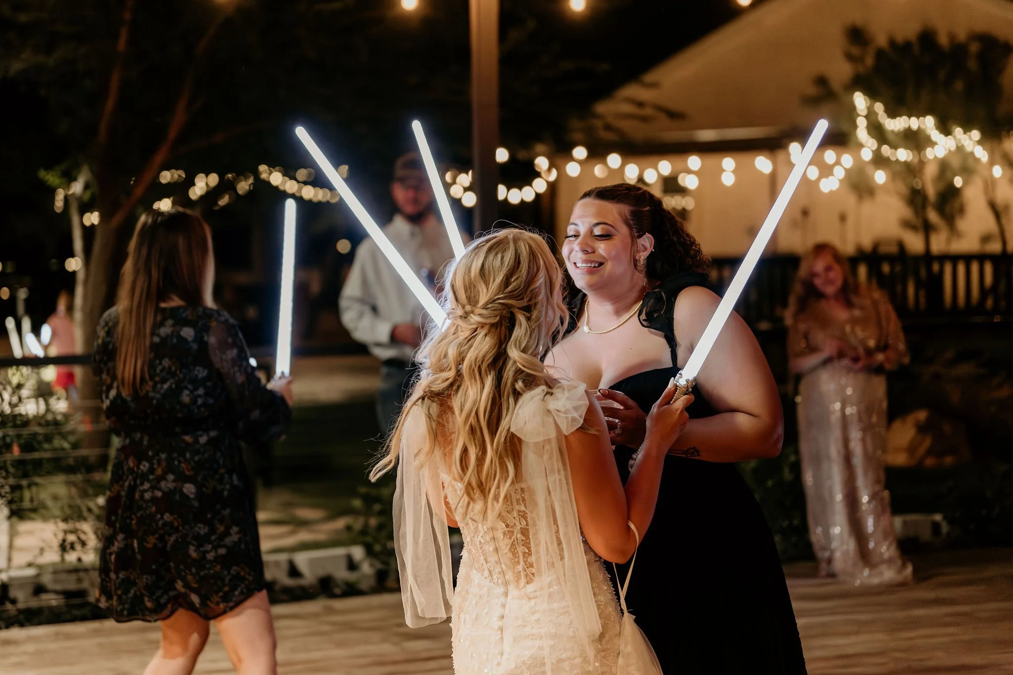 Two women dancing and smiling at a nighttime event, holding light-up sticks. One woman has long blonde hair and is wearing a light-colored dress, while the other has dark hair in a ponytail and is wearing a black dress. There are other people in the 