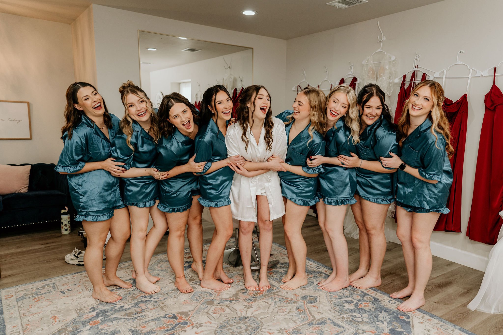 A group of nine women, including a bride in white pajamas and eight bridesmaids in matching teal satin pajamas, are laughing and smiling together in a bridal suite with red dresses on hangers in the background.
