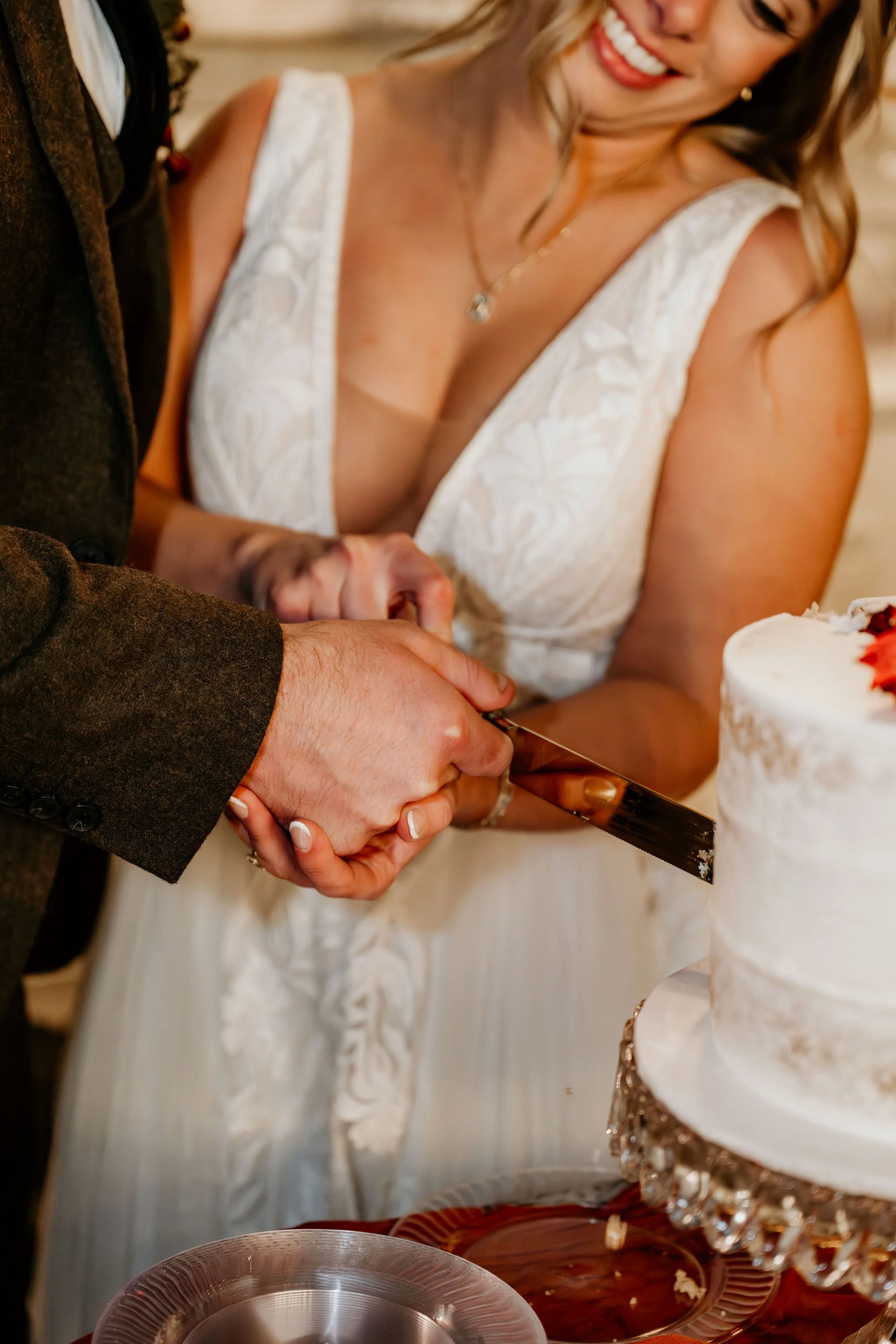 A bride and groom cut their wedding cake together, holding a cake knife. The bride is smiling and wearing a white lace dress, and the groom is in a brown suit. The wedding cake is white with decorative accents, and the scene appears to be in a celebr