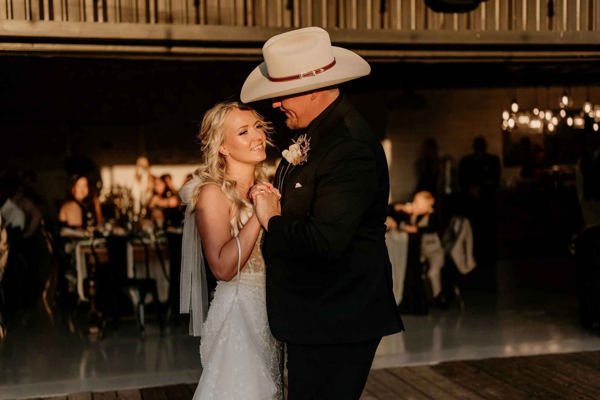 A bride and groom dancing at their wedding reception, sharing a close moment. The bride is in a white dress, smiling, with long blonde hair. The groom is wearing a black suit with a white hat and is holding the bride's hands. The background shows a d