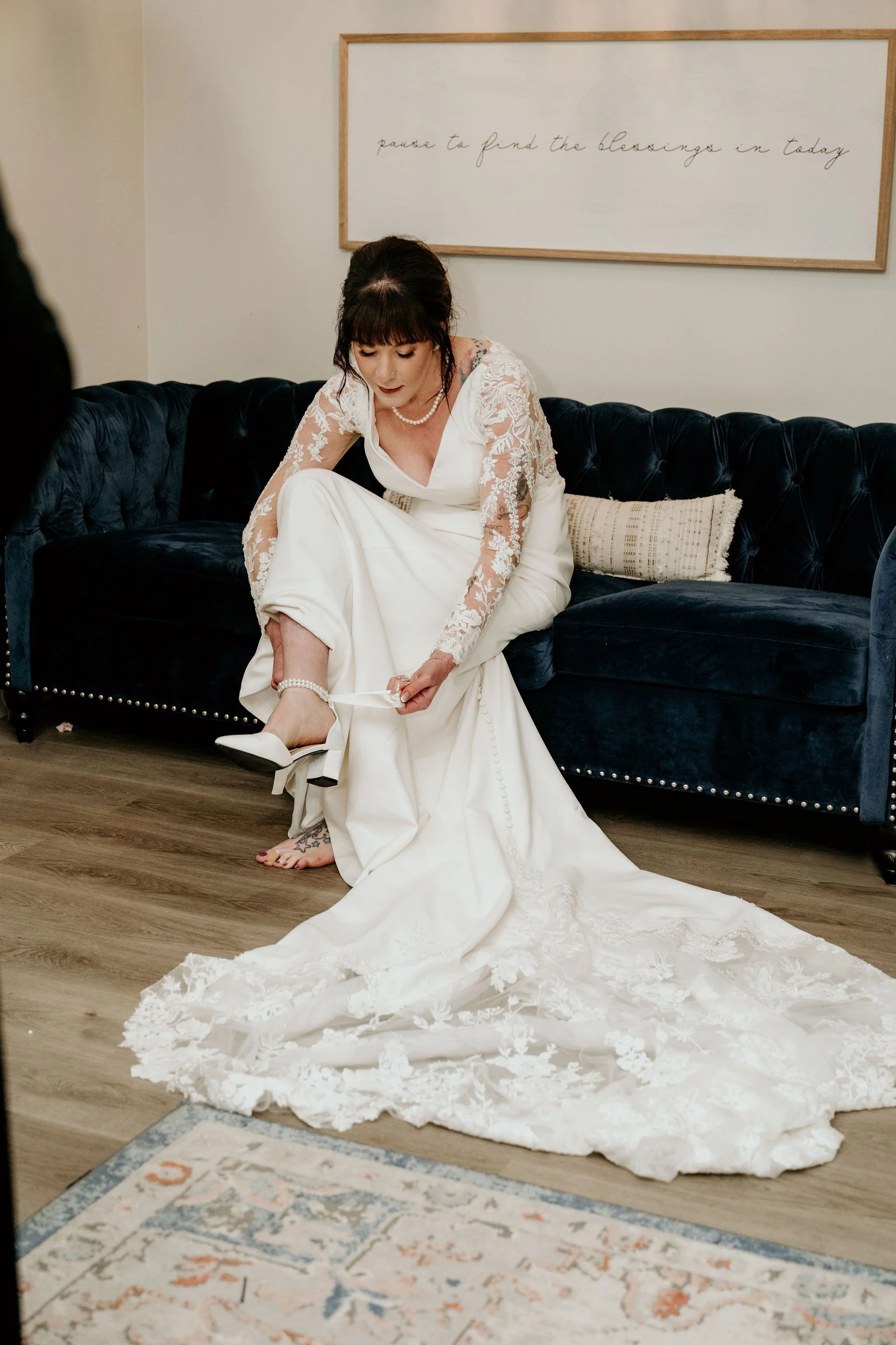 A woman in a wedding dress sits on a dark blue velvet couch, adjusting her white shoe while her other foot is bare. There is a lace train on the floor, and a framed quote on the wall behind her.