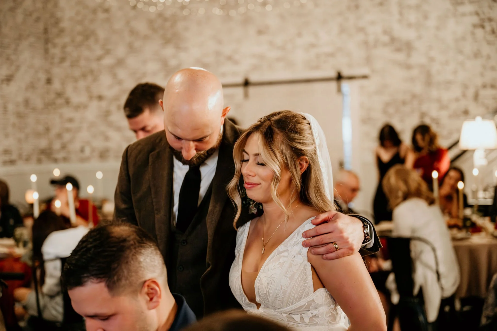 A bride and groom share a special moment with a man at their wedding reception, surrounded by guests in a rustic, warmly lit venue.