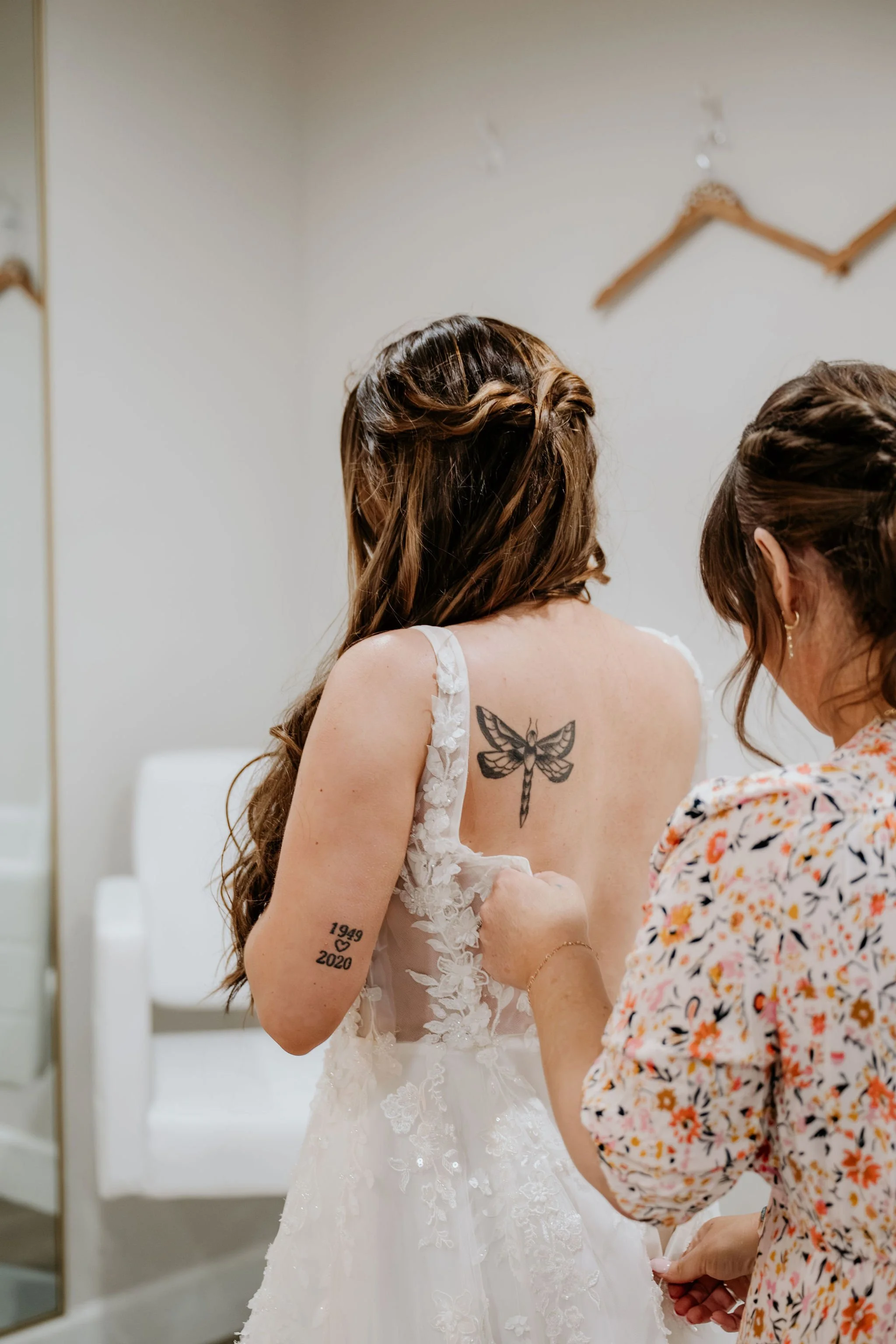 A bride in a wedding dress with floral lace details getting ready with assistance from another woman. The bride has tattoos, including a butterfly on her back and years '1949' and '2020' on her arm.