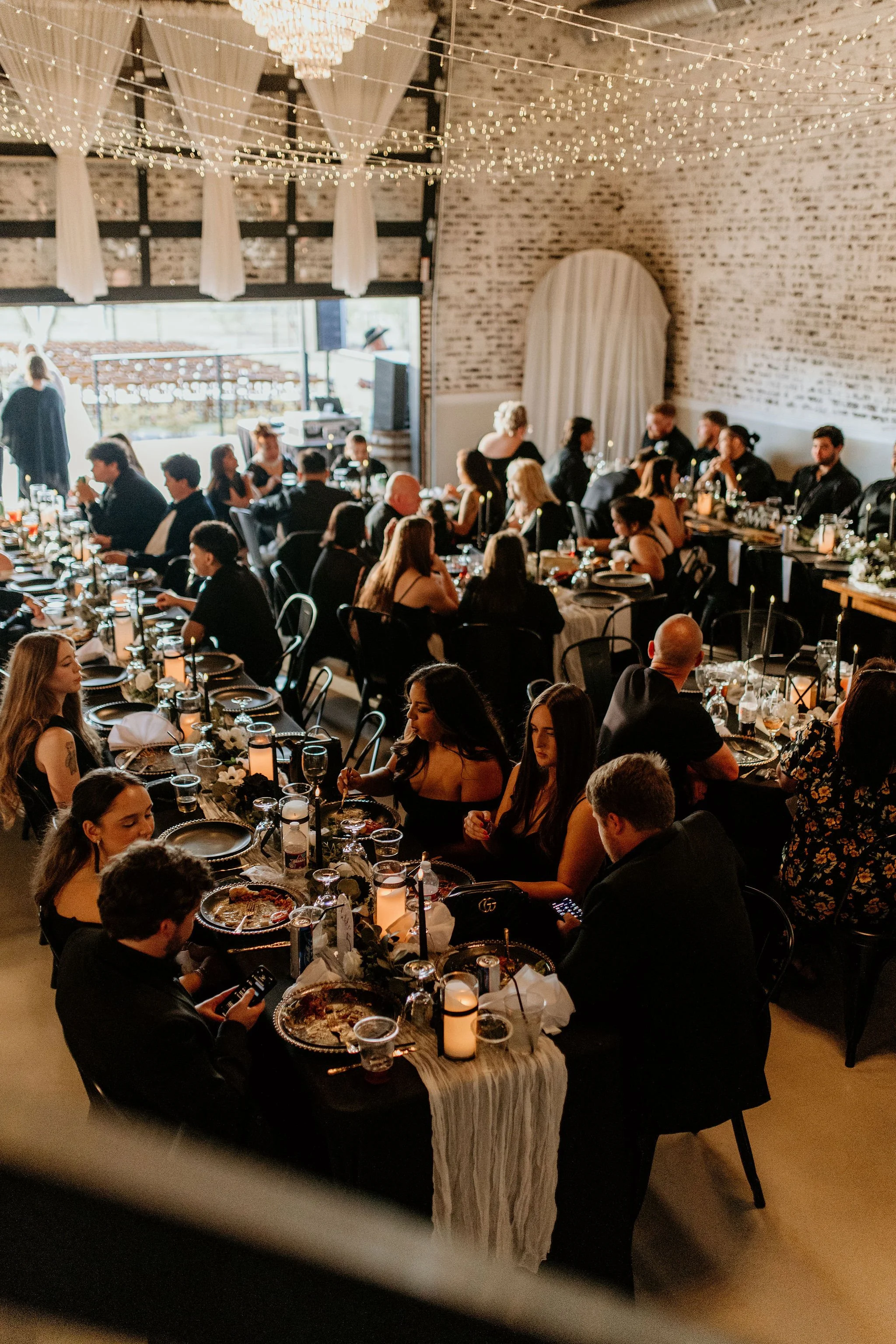 Indoor wedding reception with elegantly decorated long tables, guests in formal attire, candlelit centerpieces, string lights hanging from the ceiling, and a brick wall background.