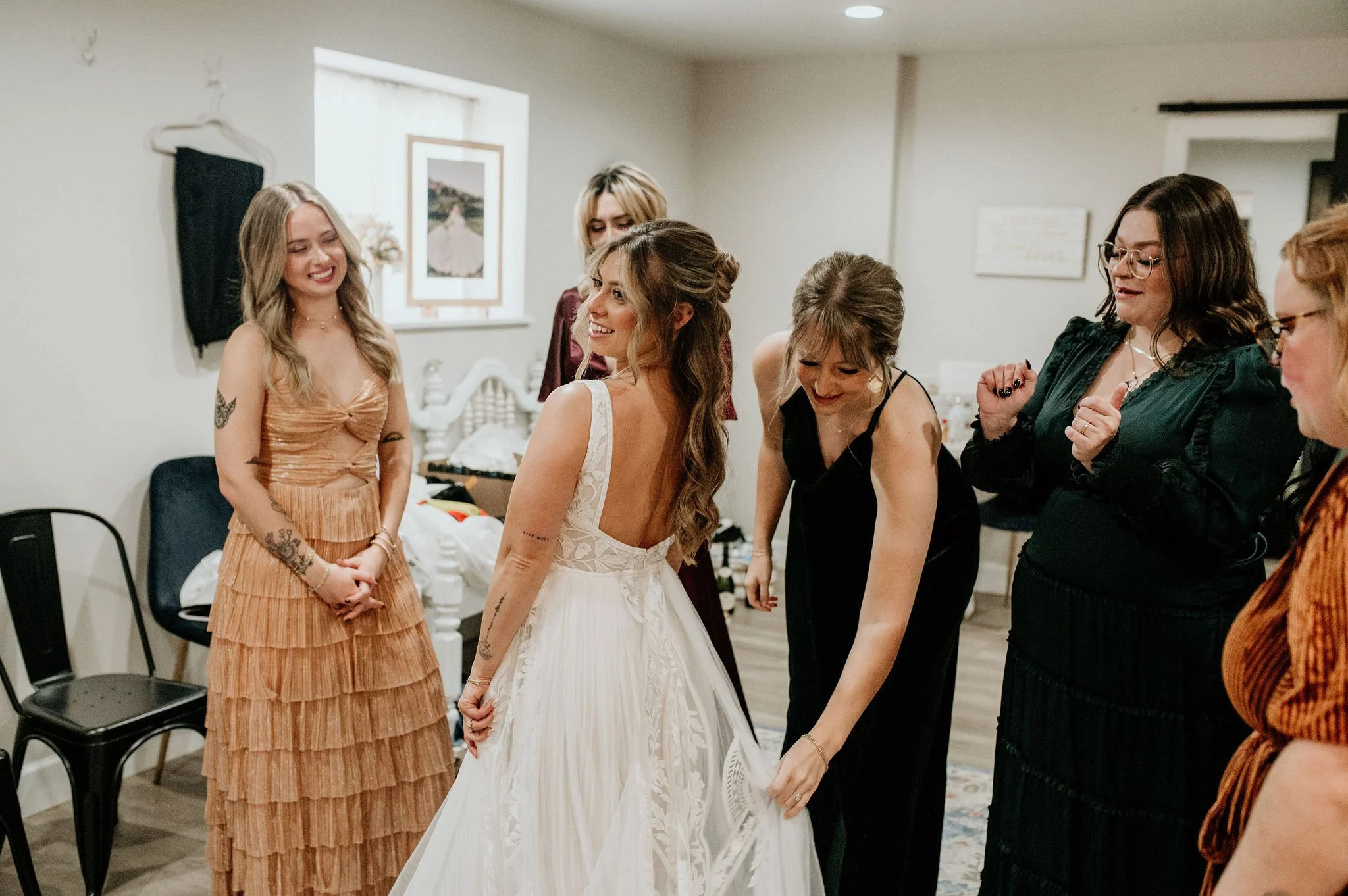 Women preparing for a wedding in a cozy room, with the bride in a white gown and friends around her adjusting her dress.