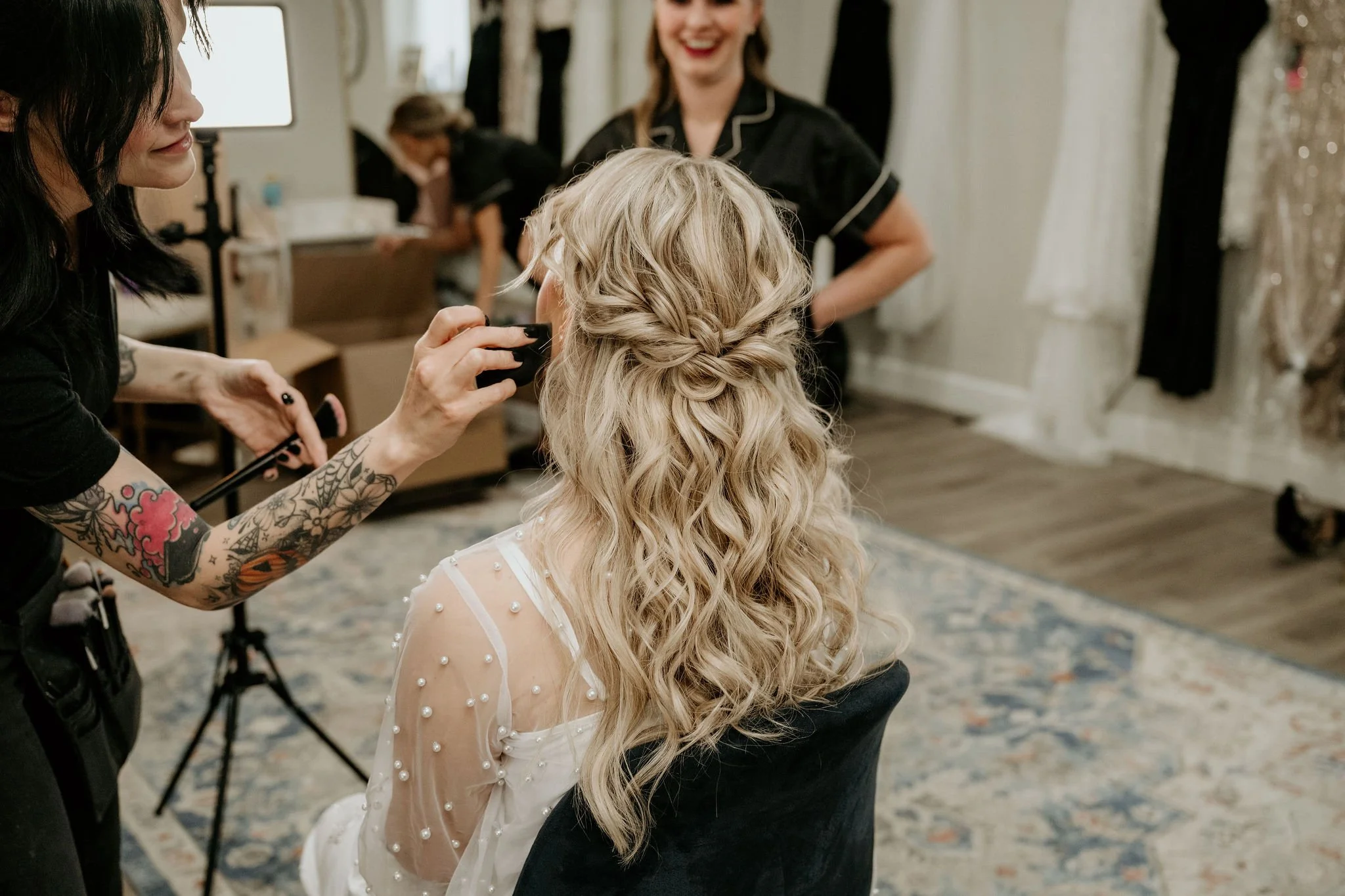 A makeup artist with tattooed arms applying makeup to a bride with long blonde hair styled in loose curls, while two other women in black outfits look on and smile, inside a bridal boutique with wedding dresses hanging in the background.