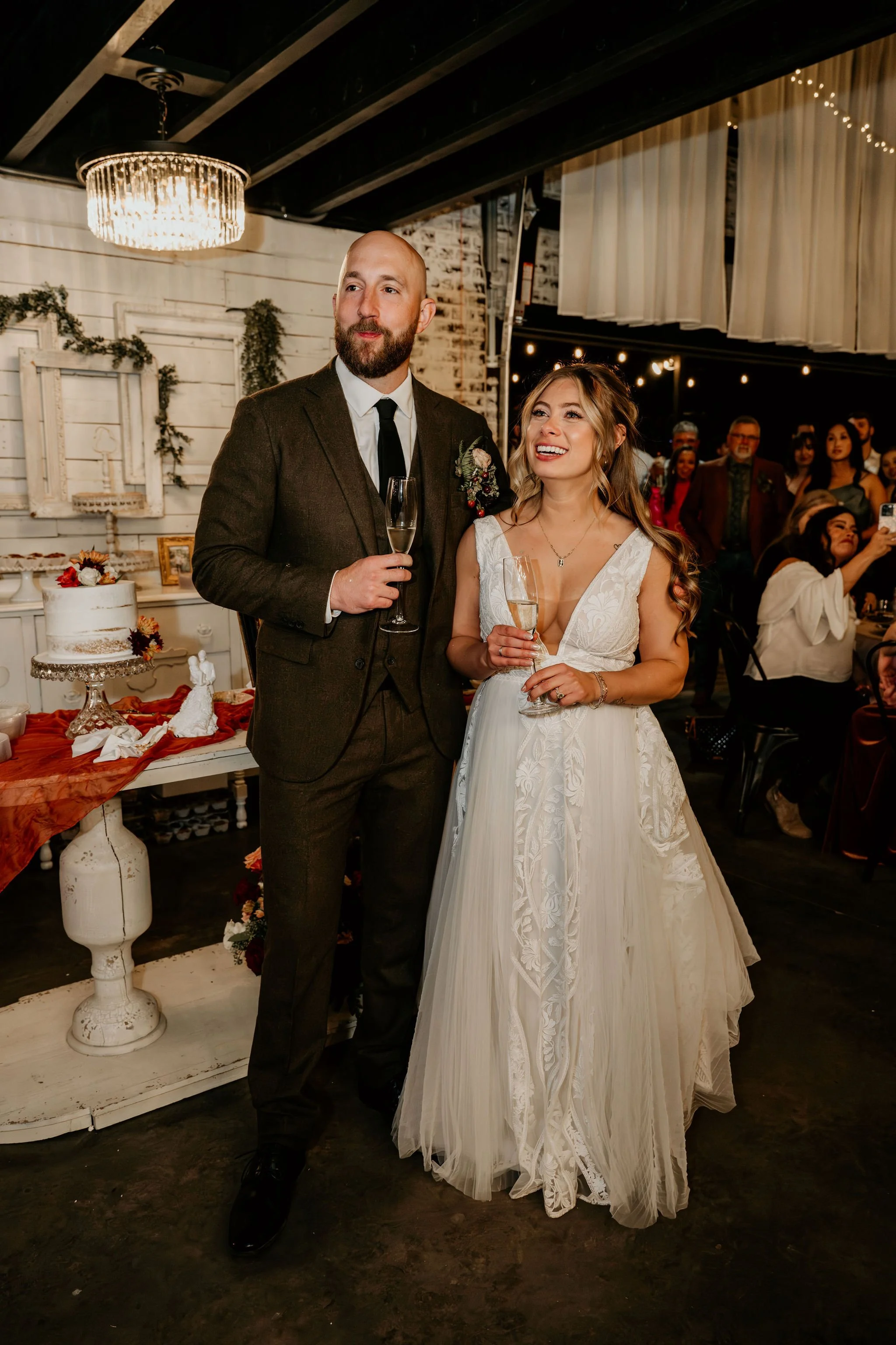 A bride and groom standing together at their wedding reception, both holding champagne glasses, surrounded by guests and wedding decorations.