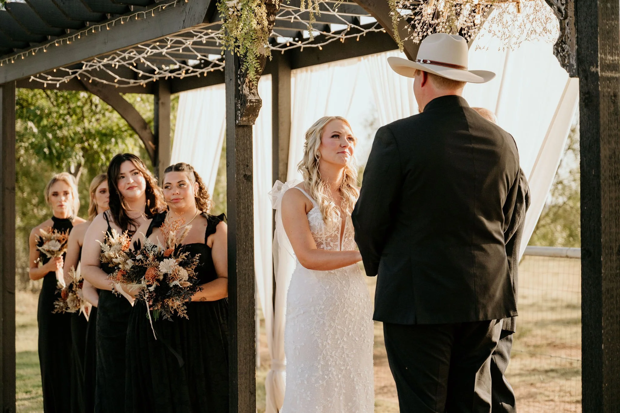 Bride and groom exchanging vows during outdoor wedding ceremony, with bridesmaids and attendants watching in the background.