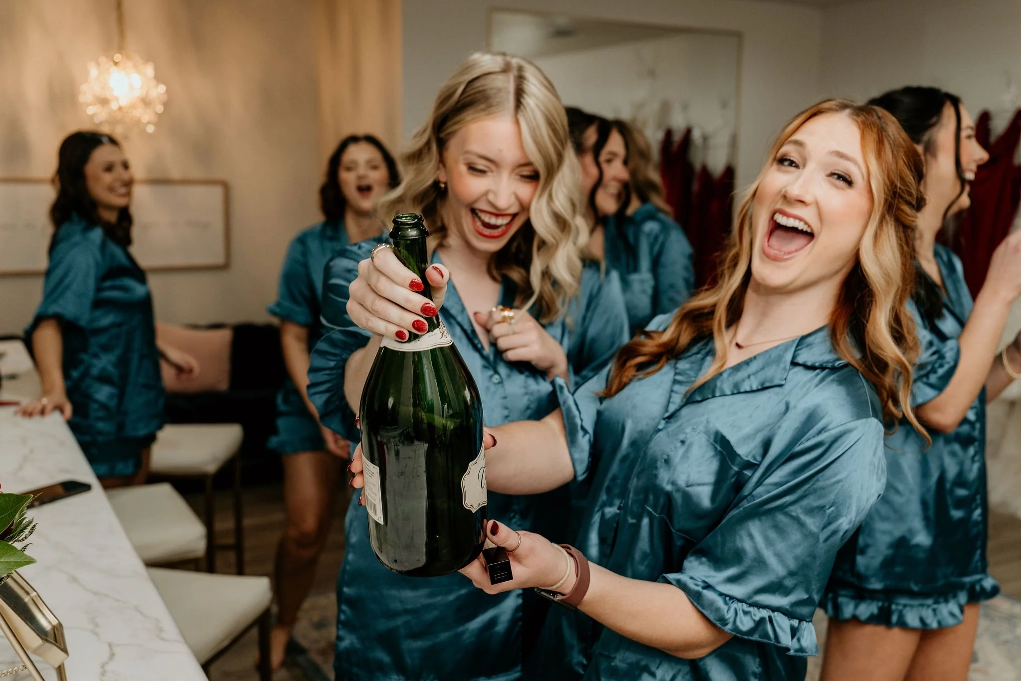 Group of women in matching blue satin pajamas celebrating and opening a bottle of champagne in a hotel room.