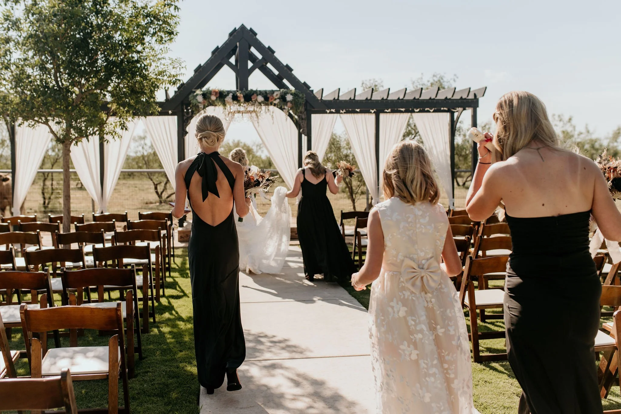 Women attending an outdoor wedding ceremony with a decorated altar, bridesmaid in black gown, flower girl in white dress, and wedding dress carried down the aisle.