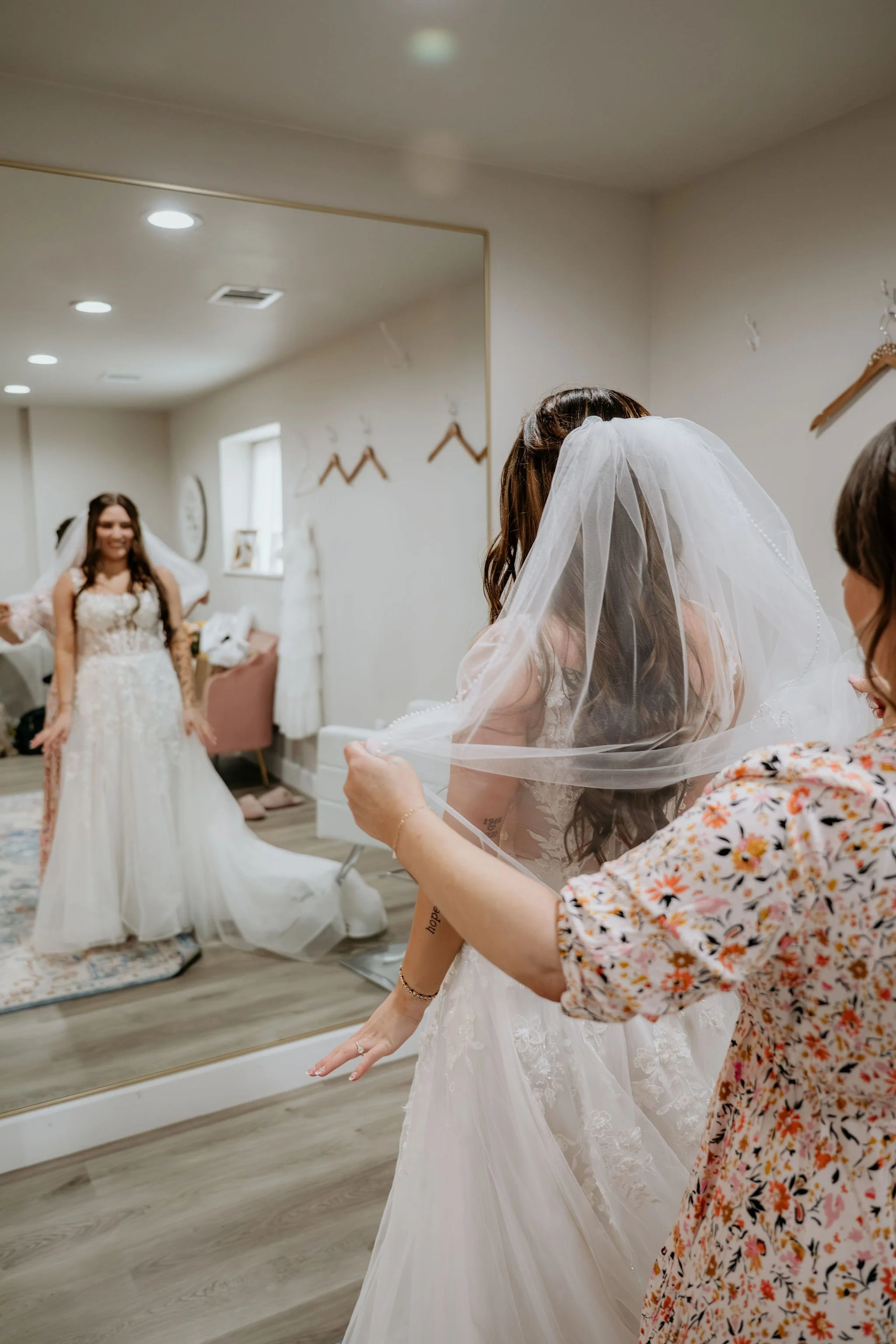 Bride trying on wedding dress with helper adjusting veil, mirror reflecting excited bride in wedding gown.