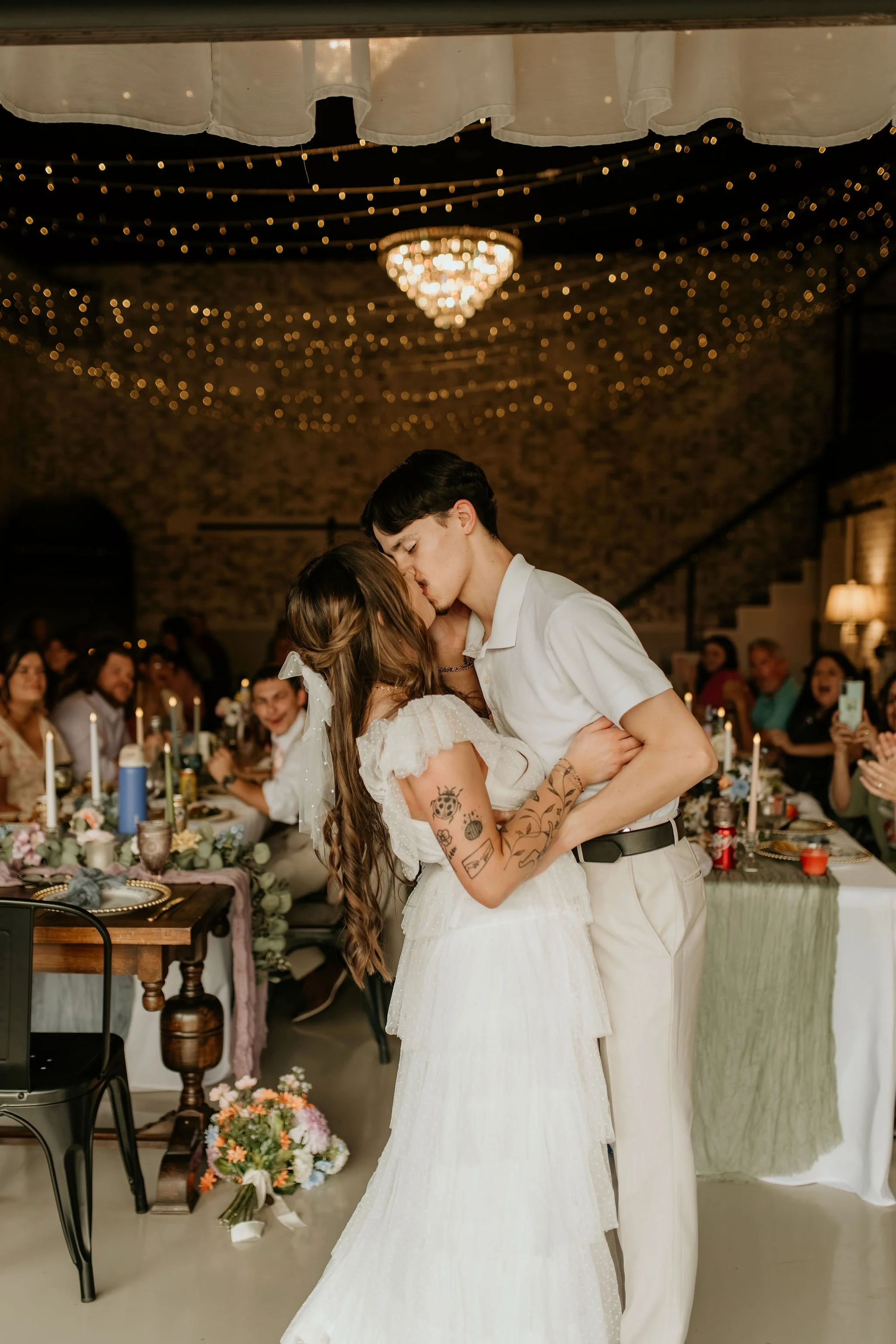 A couple sharing a kiss at a wedding reception, with guests in the background sitting at a decorated table under string lights and a chandelier.
