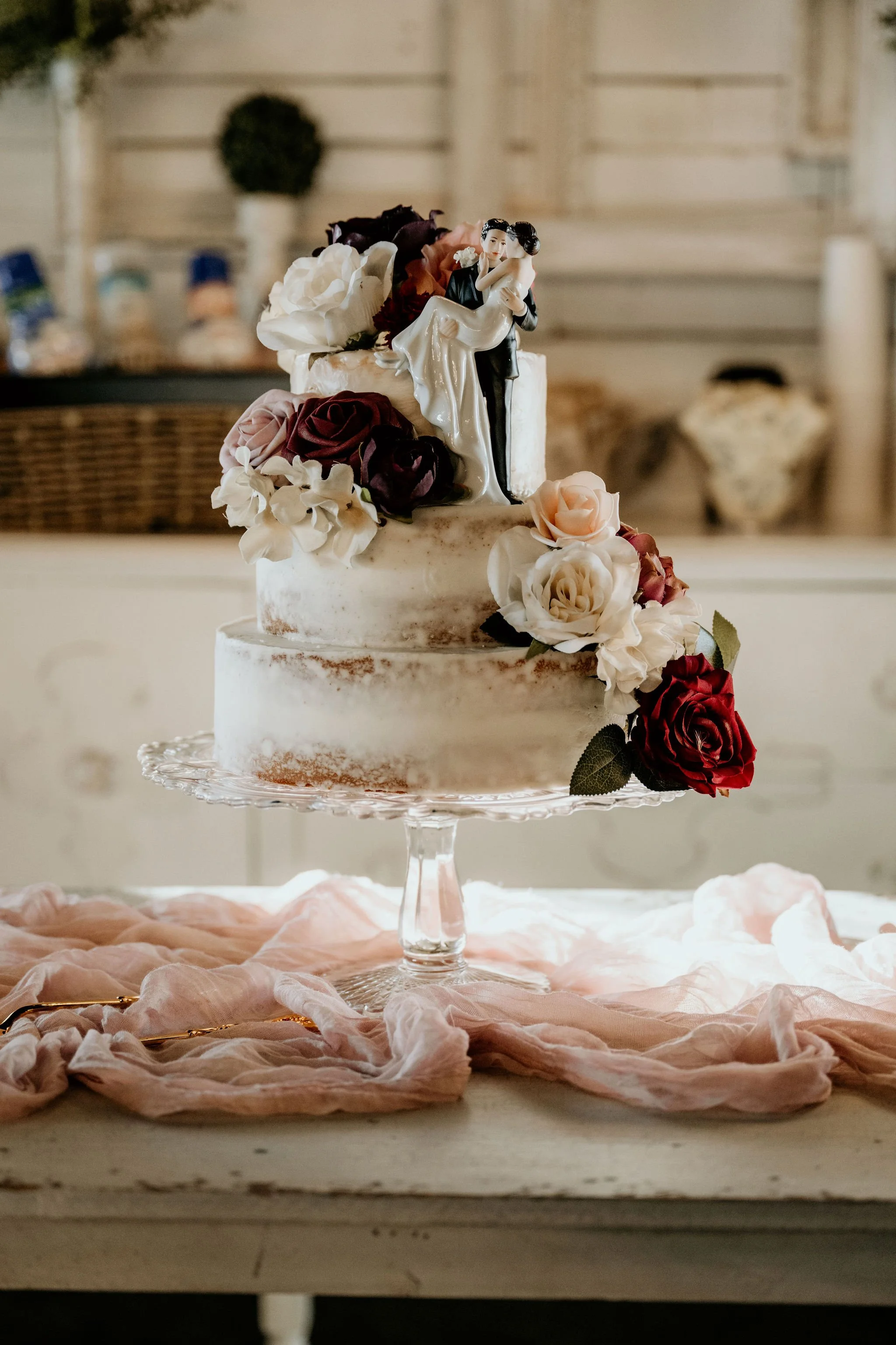 A wedding cake with white frosting, decorated with purple, white, and peach roses, topped with a figurine of a bride and groom, on a glass cake stand, with pink fabric underneath and blurred background.