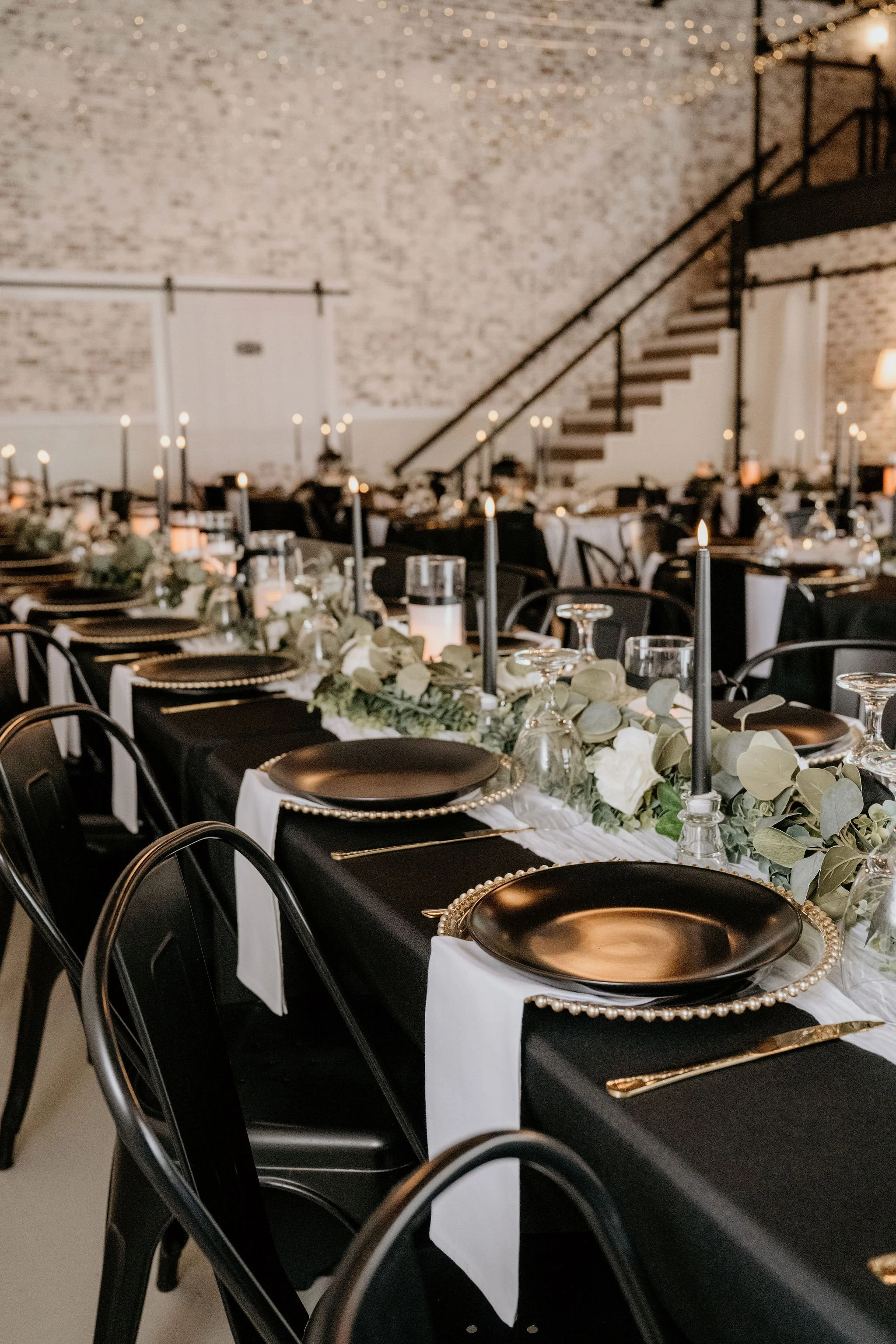 Elegant banquet table with black and white color scheme, black plates with pearl edges, gold cutlery, black candles, floral centerpiece with white flowers and green leaves, set in a rustic industrial venue with exposed brick walls, string lights, and