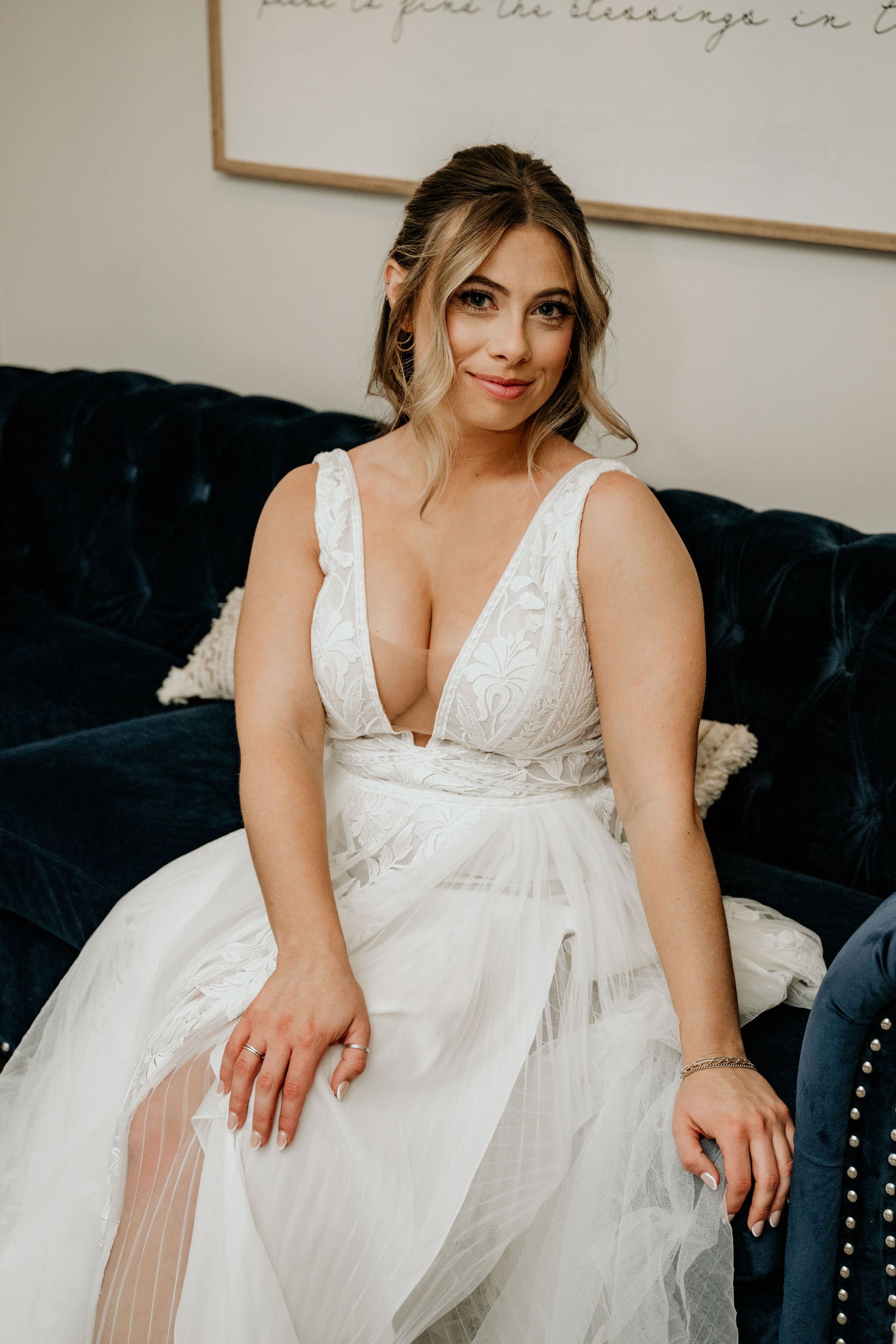 A woman in a white wedding dress sitting on a dark velvet couch, smiling at the camera.