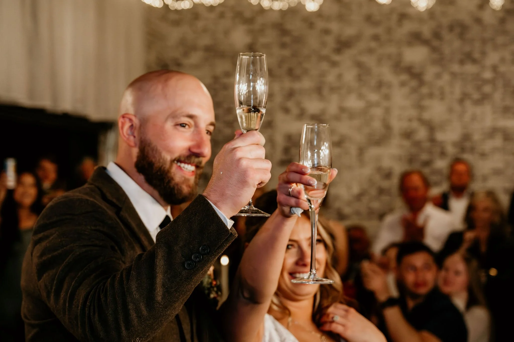 A man and a woman raise champagne glasses in a toast at a celebration, surrounded by a lively crowd in an indoor setting.