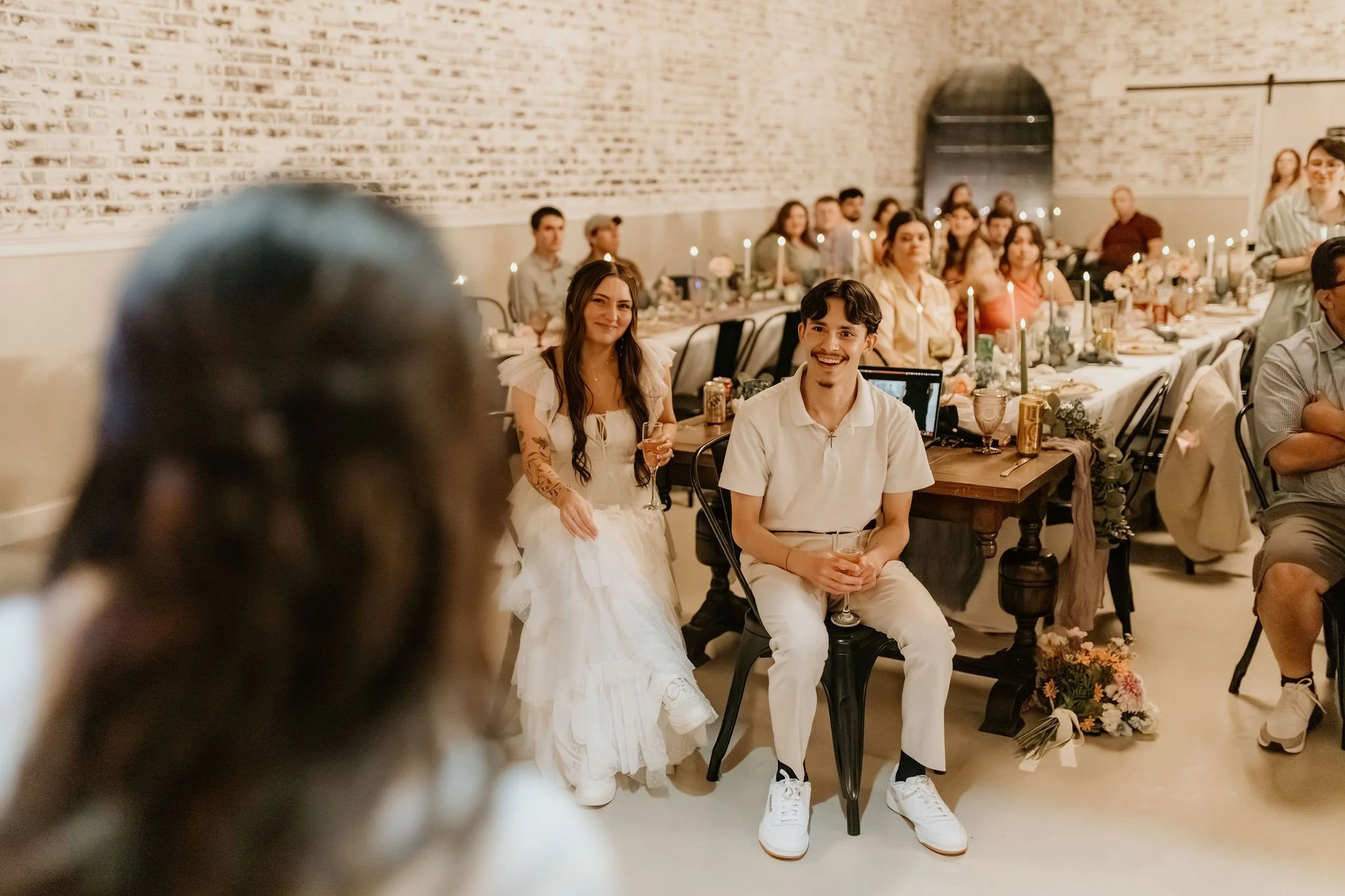 A wedding reception with guests seated at long decorated tables with candles and flowers, focusing on a smiling woman in a wedding dress and a man in white attire sitting and holding drinks.