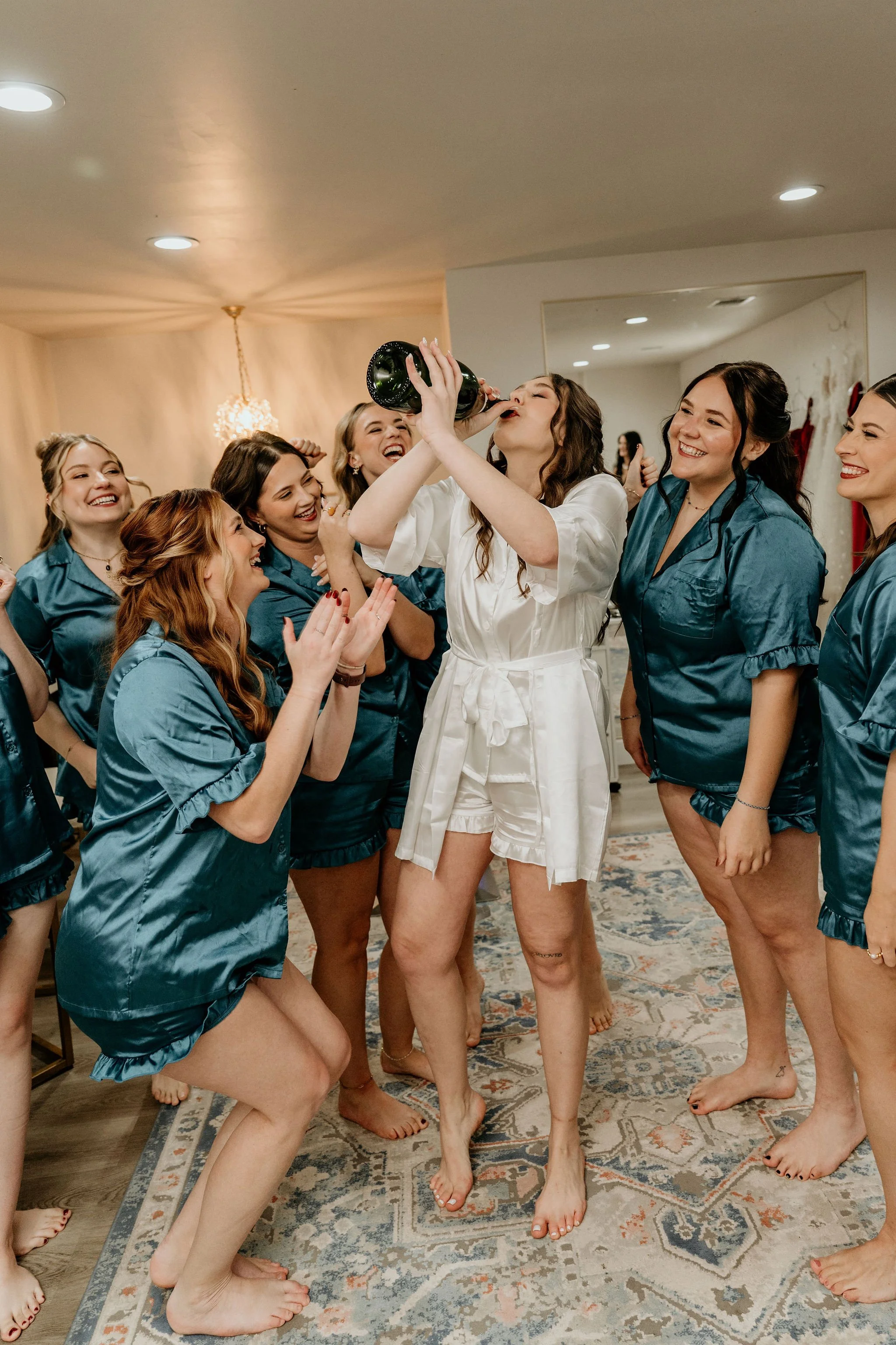 A bride wearing white pajamas is drinking champagne from a bottle surrounded by women in matching teal pajamas, celebrating.