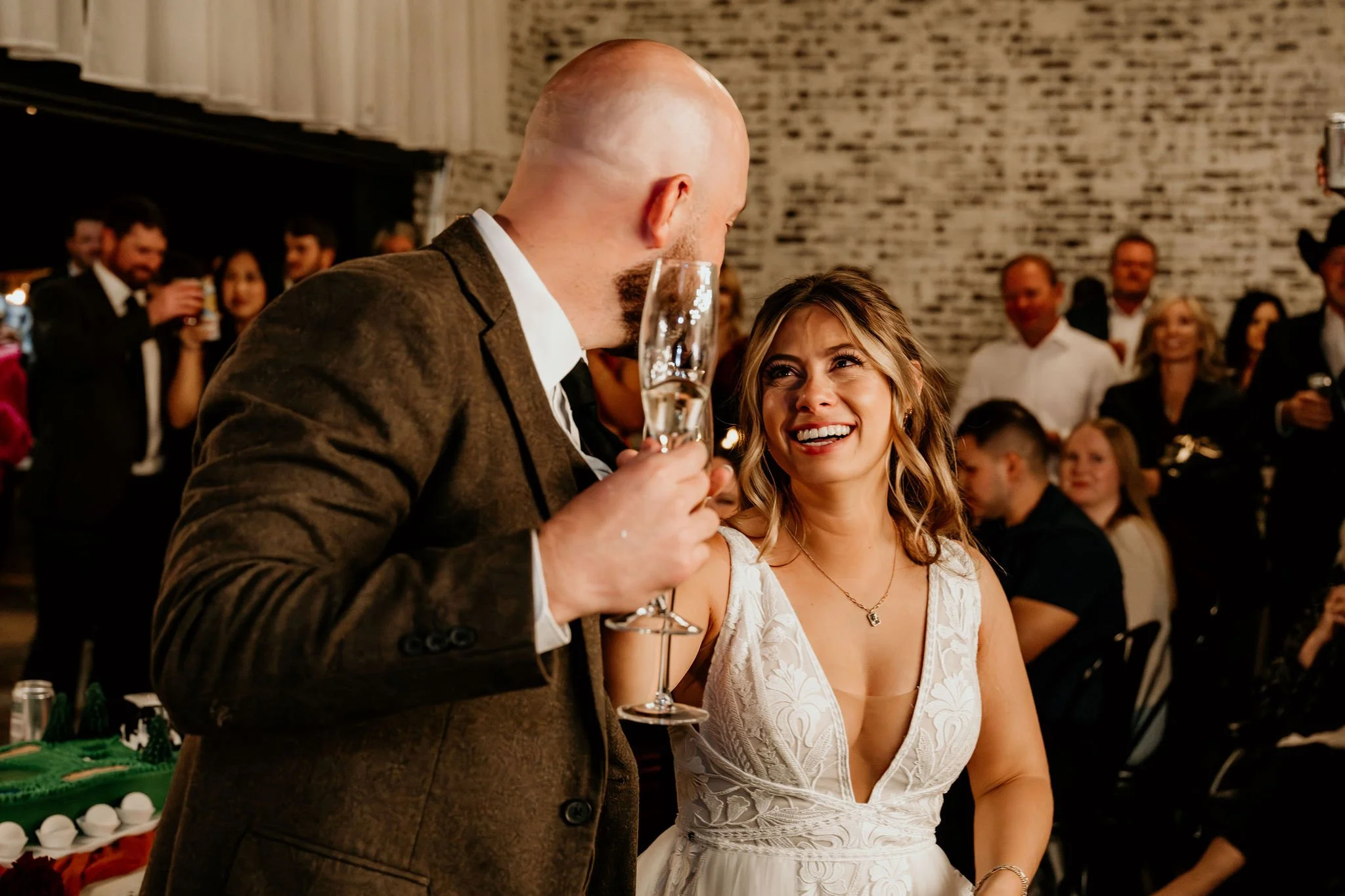 A man in a brown suit holding a champagne glass while talking to a smiling woman in a white wedding dress at a celebration.