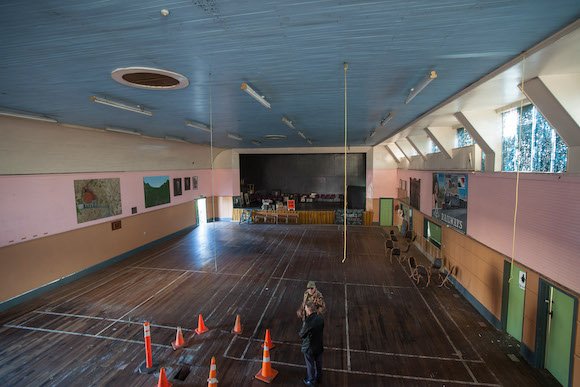 Empty gymnasium with a person standing near orange cones, wooden floor, painted walls, and stage at the front.