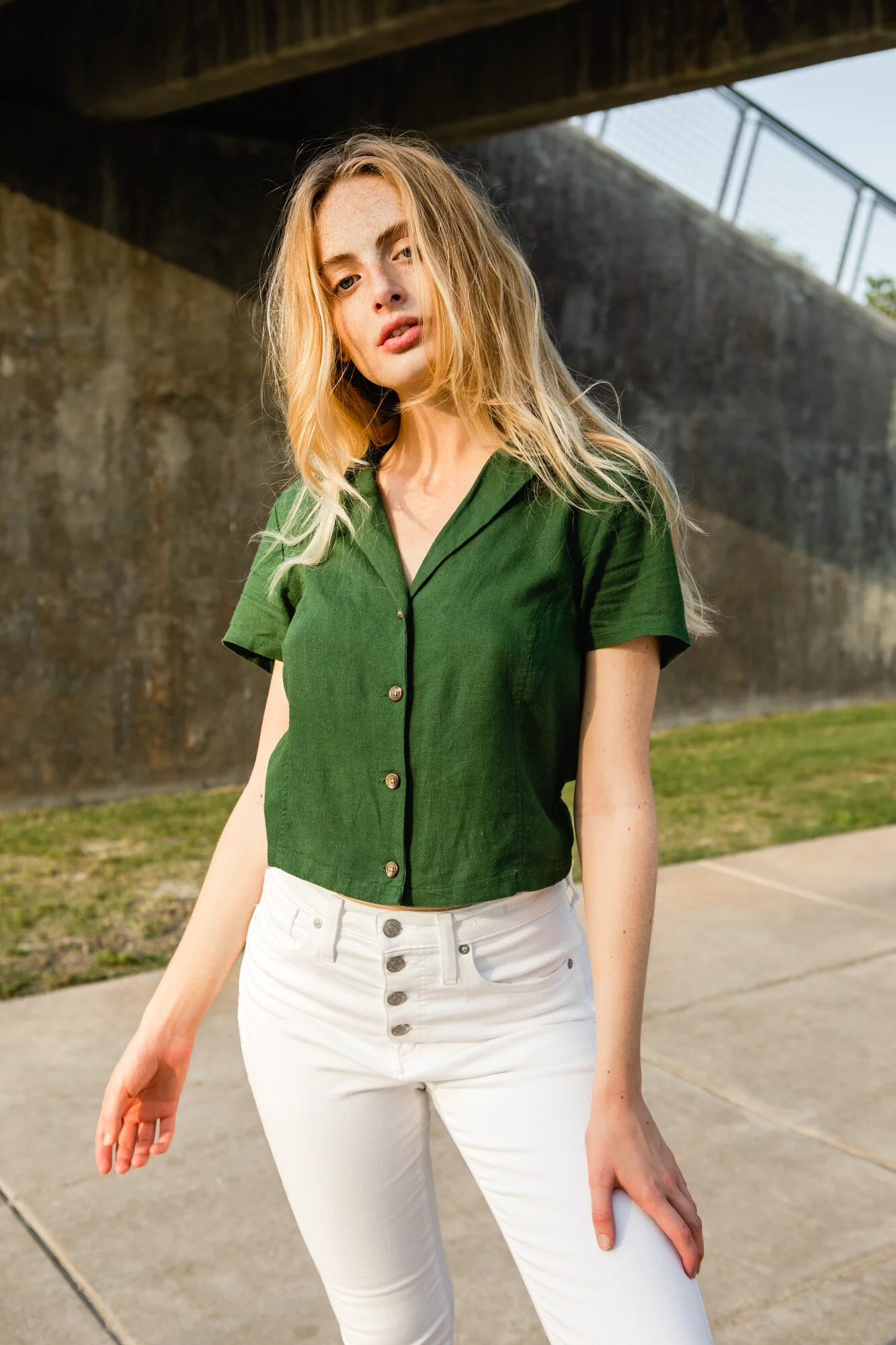 A young woman with long, wavy blonde hair wearing a green short-sleeved button-up shirt and white high-waisted pants standing outdoors near a concrete wall and under an overpass.