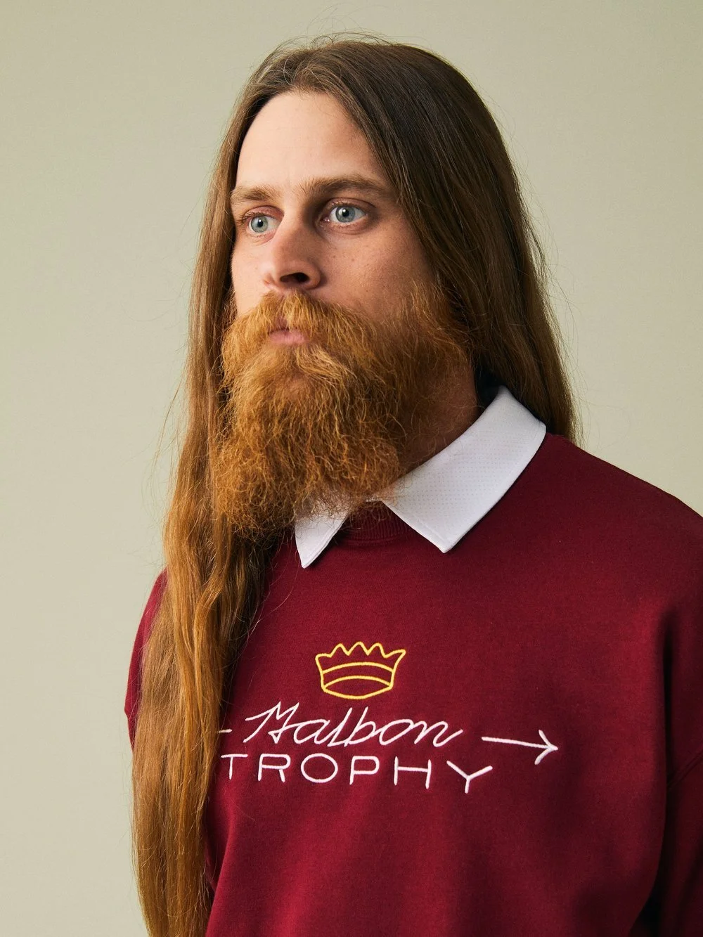 A man with long red hair and a full beard wearing a maroon sweater with white text and a yellow crown graphic, and a white collared shirt underneath.