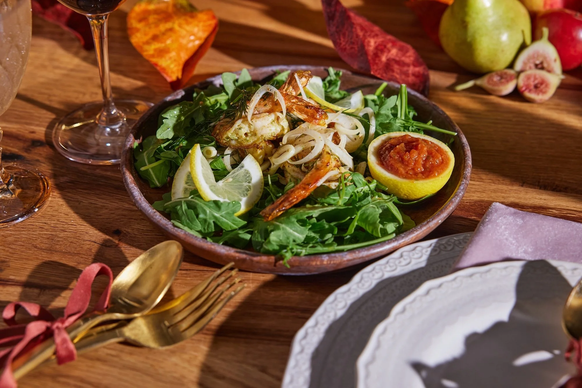 A wooden table set with a plate of salad featuring greens, grilled shrimp, lemon slices, and an orange-colored sauce in a lemon half, surrounded by wine glasses, gold utensils with pink ribbons, decorative leaves, and figs in the background.