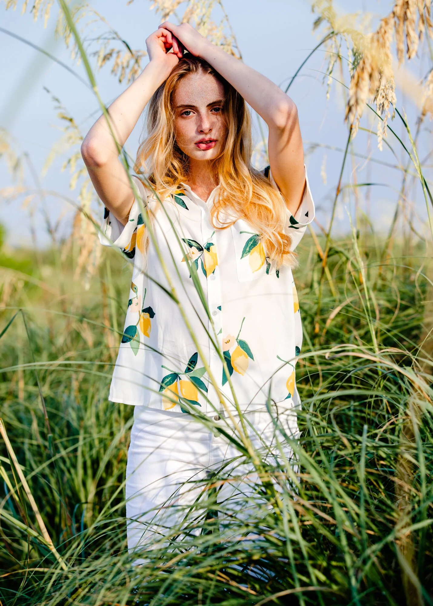 A woman with long, wavy blonde hair standing among tall grass, wearing a white shirt with a lemon pattern and white pants, with her arms raised above her head.