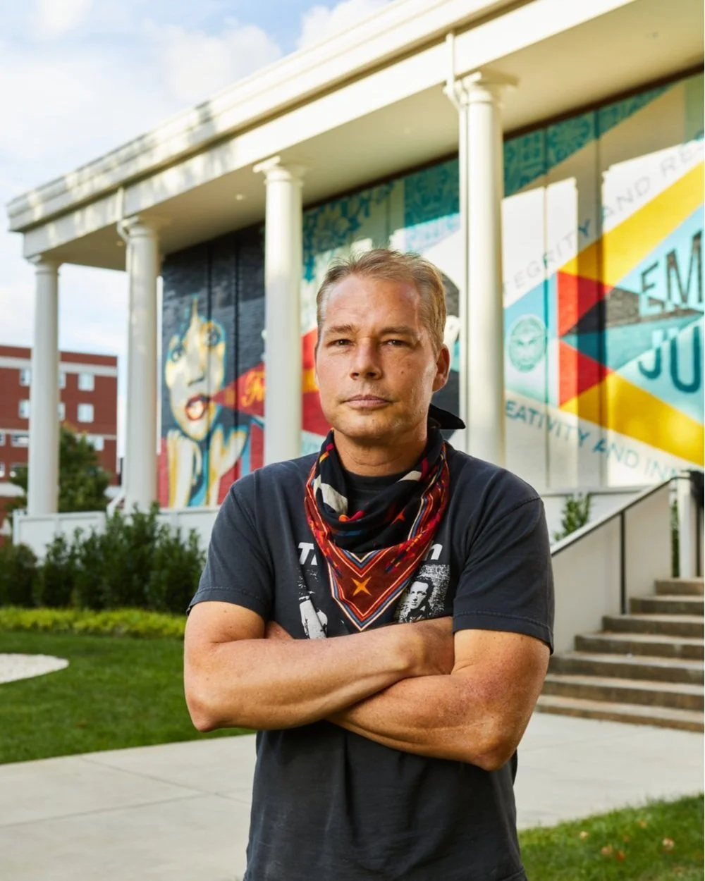 A man with crossed arms standing outside a building with colorful murals on the wall behind him.