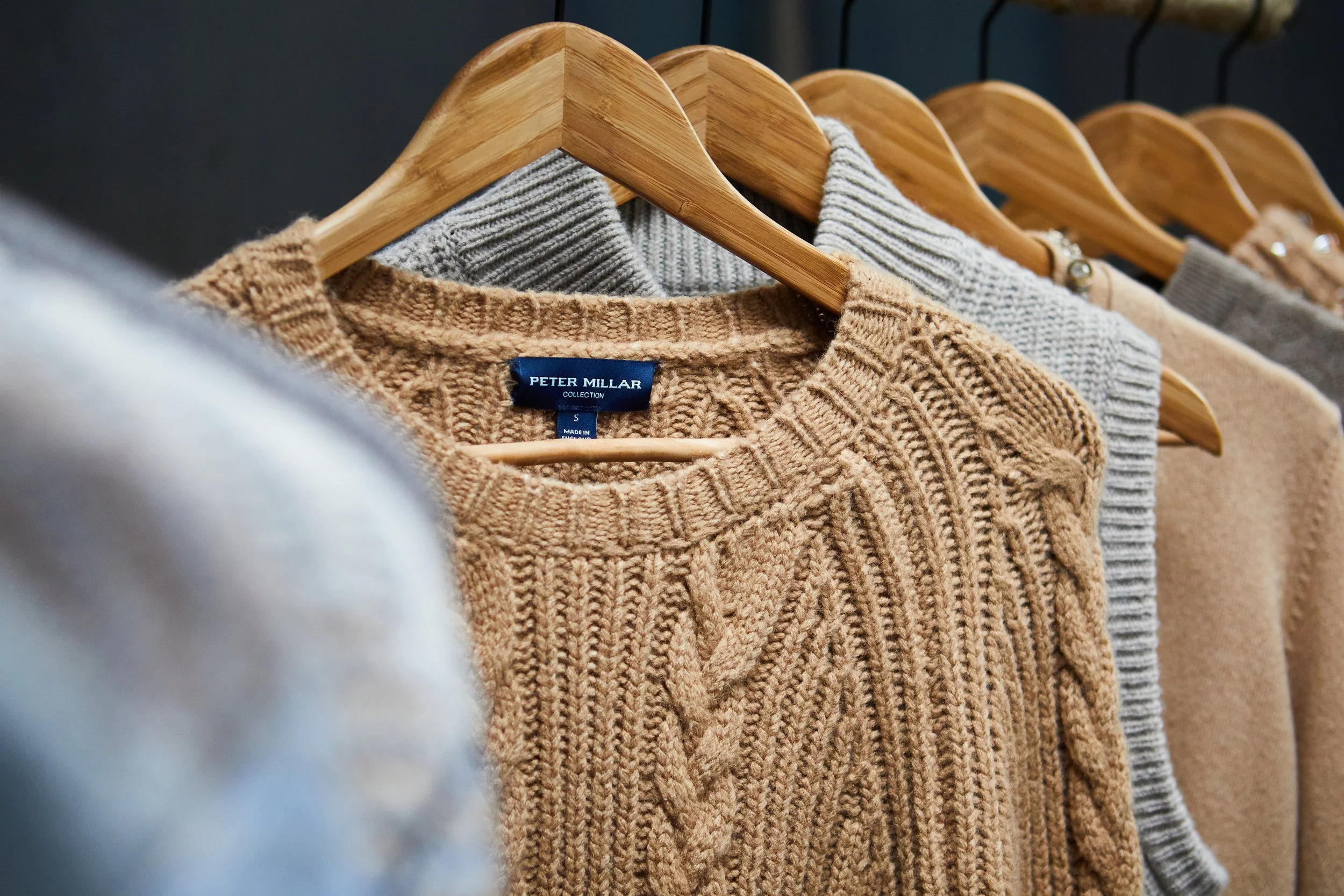 A row of knit sweaters in neutral colors hanging on wooden hangers, with the focus on a tan cable knit sweater from the Peter Millar collection.
