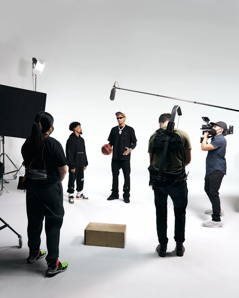 A film crew filming a basketball player in a studio with white background. The player holds a basketball and wears sunglasses and jewelry, while the crew uses cameras and lighting equipment.