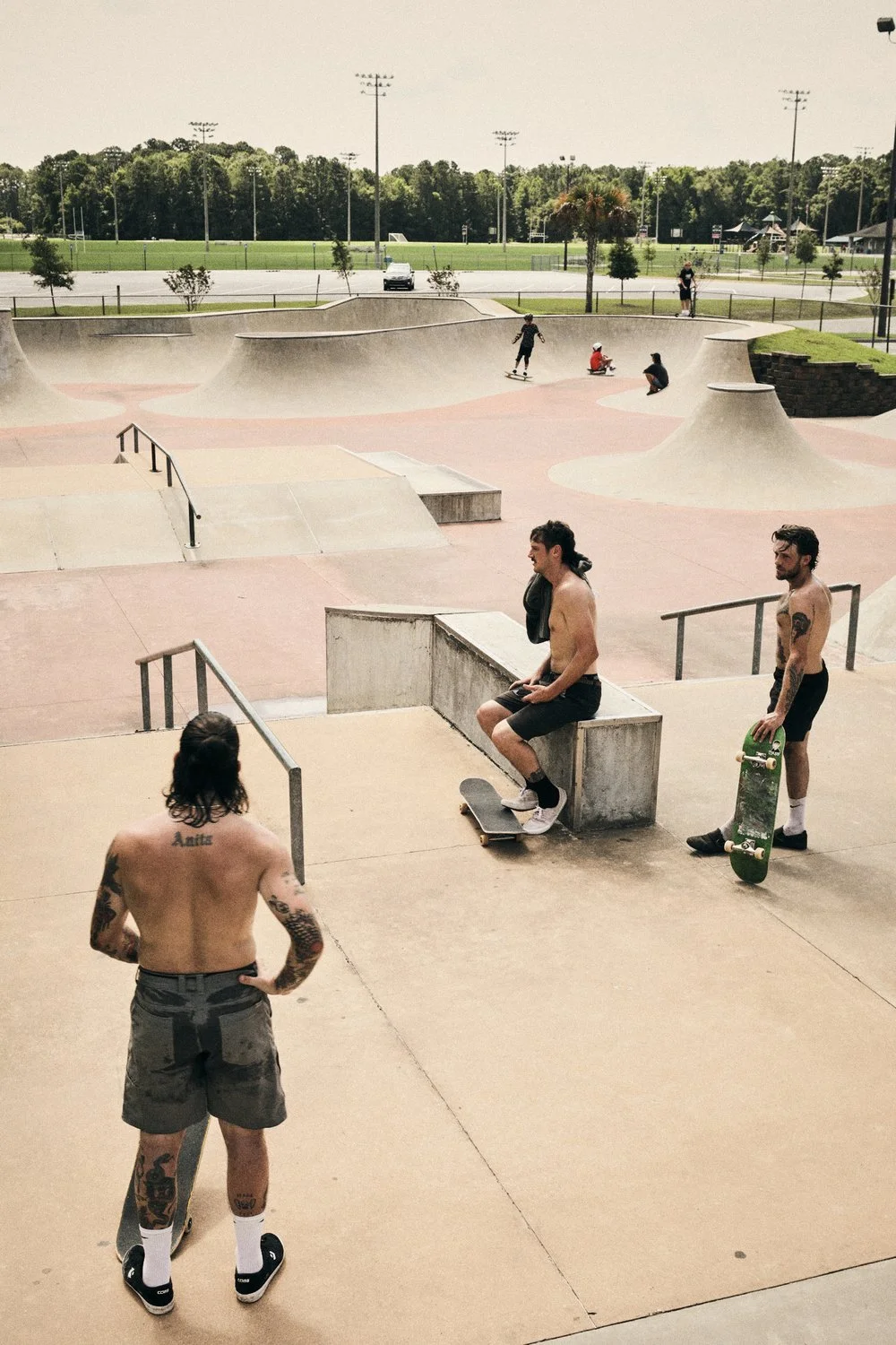 Skateboarders at a concrete skatepark with ramps and bowls, some sitting and some skating, during daytime, with a grassy field and trees in the background.