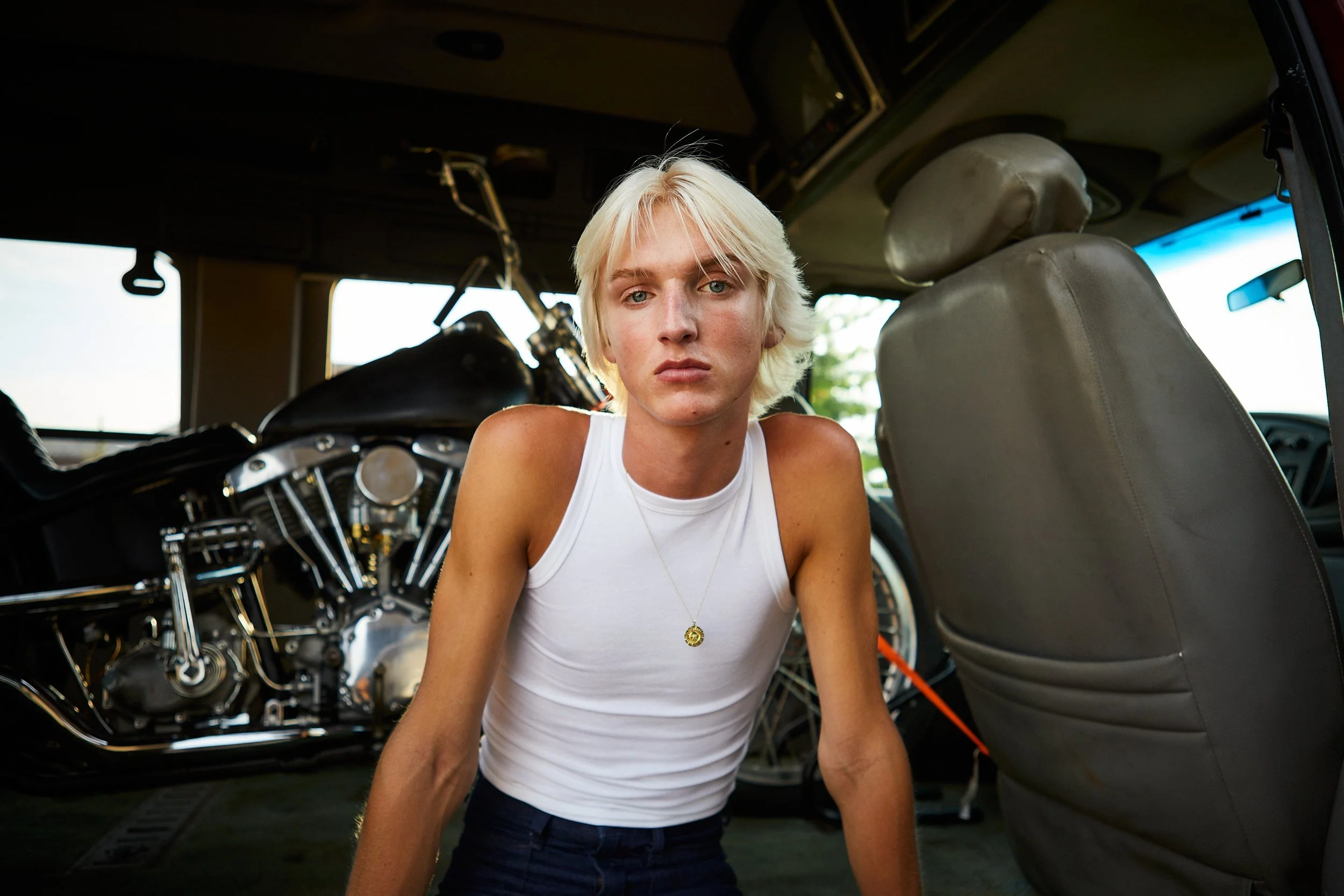 Person with short blonde hair wearing a white tank top and a gold necklace sitting inside a vehicle with a motorcycle in the background.