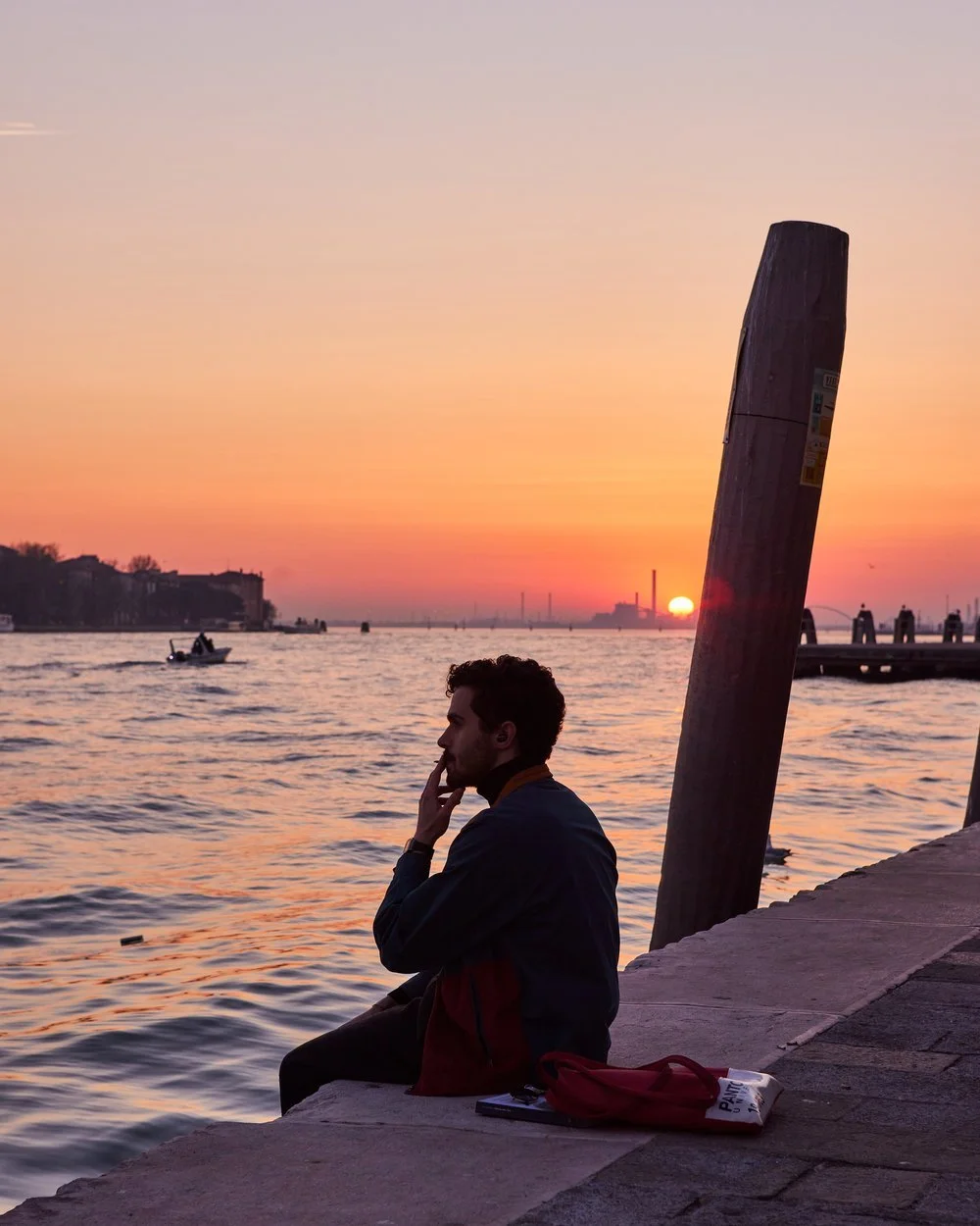 A man sitting by the water at sunset, with a boat in the background and a partially visible building on the horizon.
