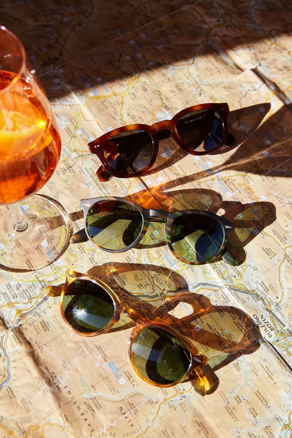 Three pairs of sunglasses and two drinks placed on a topographical map. The sunglasses are different styles and colors, casting shadows on the map.