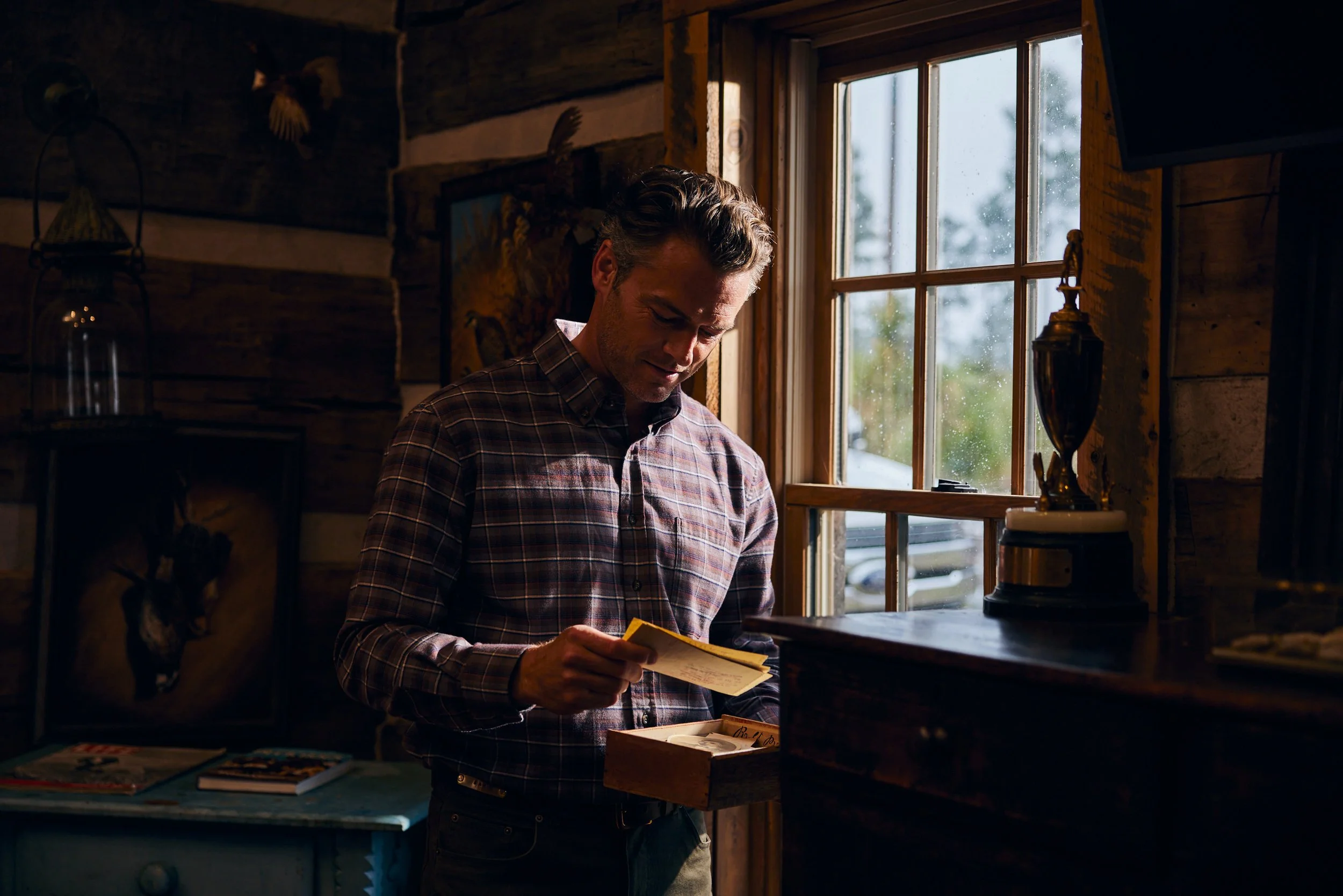 A man in a checkered shirt looking at a yellow note in a wooden box inside a rustic cabin.