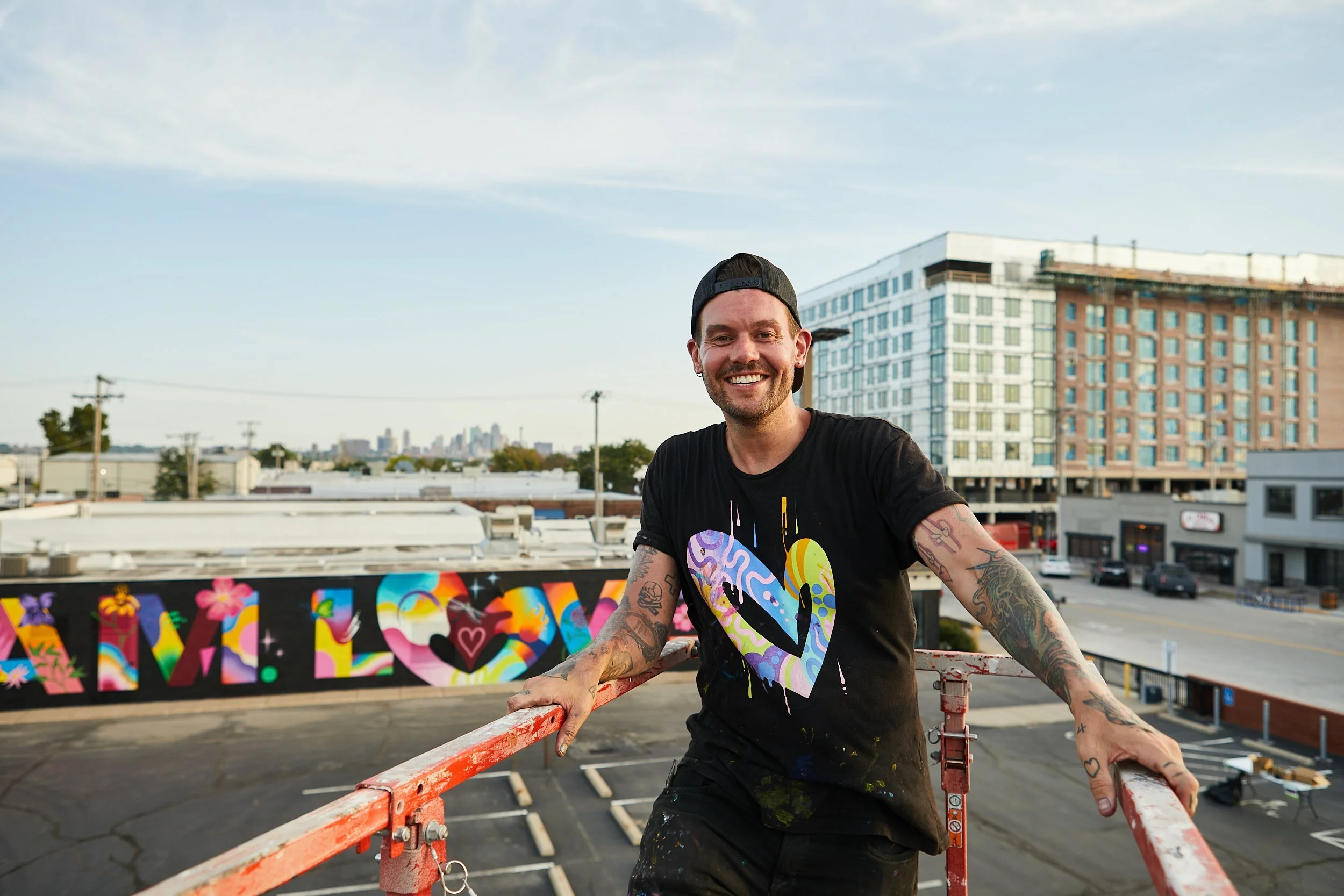 A smiling man with tattoos on his arms wearing a black t-shirt with multicolored heart design, standing on a red scaffold on a rooftop with a mural and city buildings in the background.