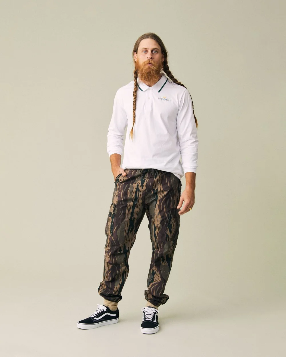 A man with long hair and a beard, wearing a white polo shirt, camouflage pants, and black sneakers, standing against a plain background.