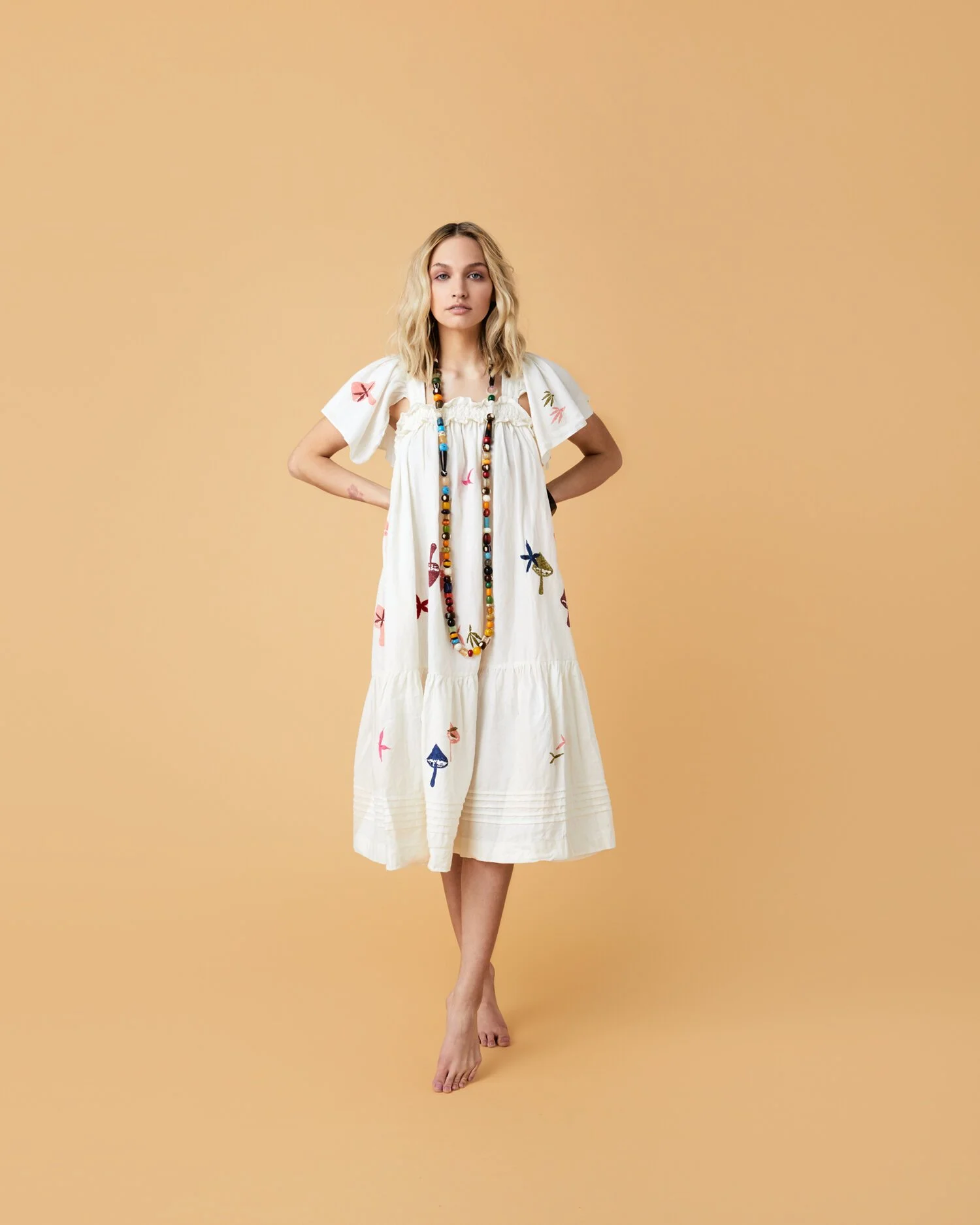 A woman in a white dress with colorful butterfly and insect embroidery, wearing a long beaded necklace, standing against a plain peach background.