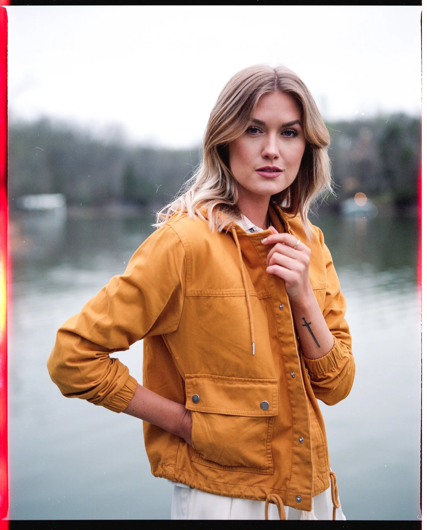 A woman with shoulder-length blonde hair wearing a mustard yellow jacket and light-colored pants standing outdoors near a body of water with trees in the background.