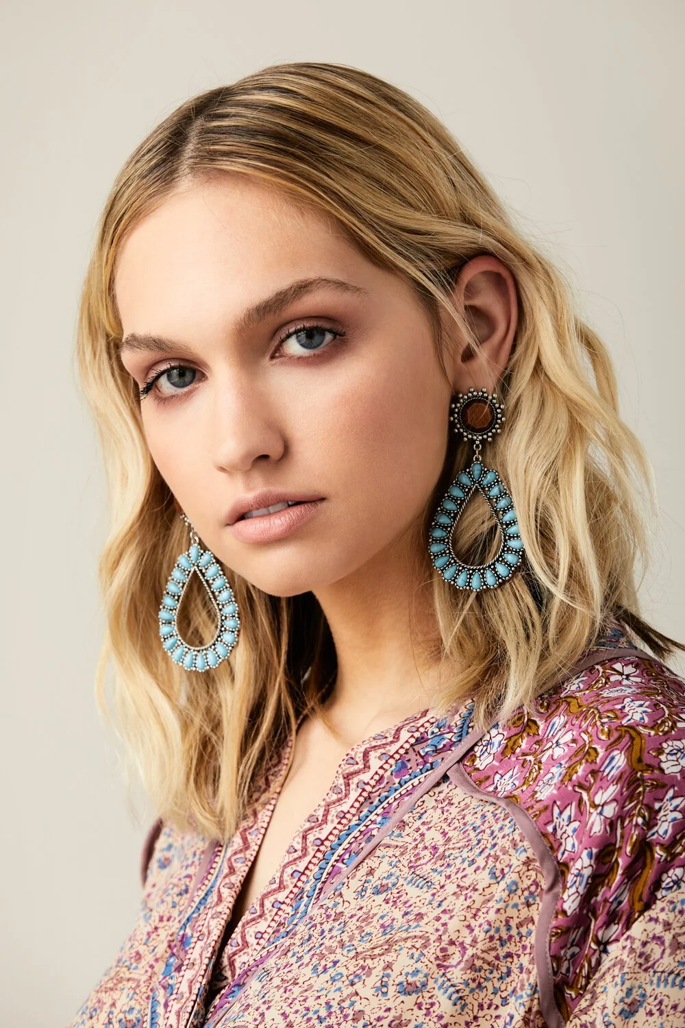 A woman with blonde hair, blue eyes, and fair skin wearing large teardrop-shaped turquoise and black earrings and a patterned blouse.