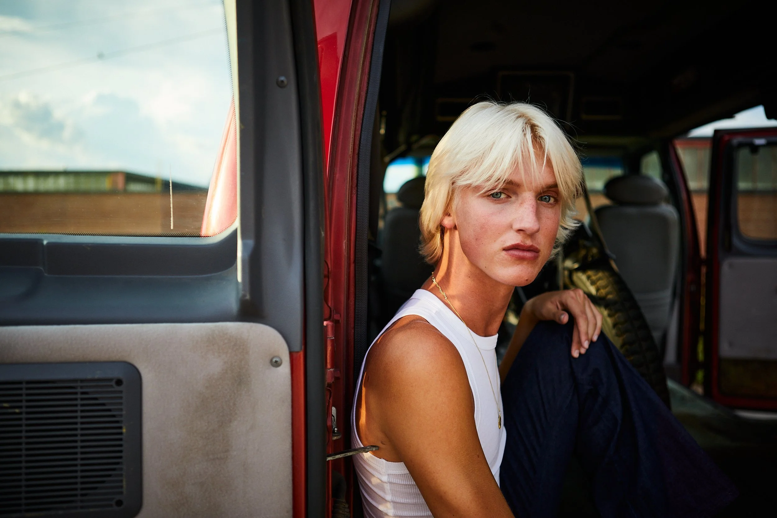 Young woman with short blonde hair sitting inside a red vehicle, looking at the camera.