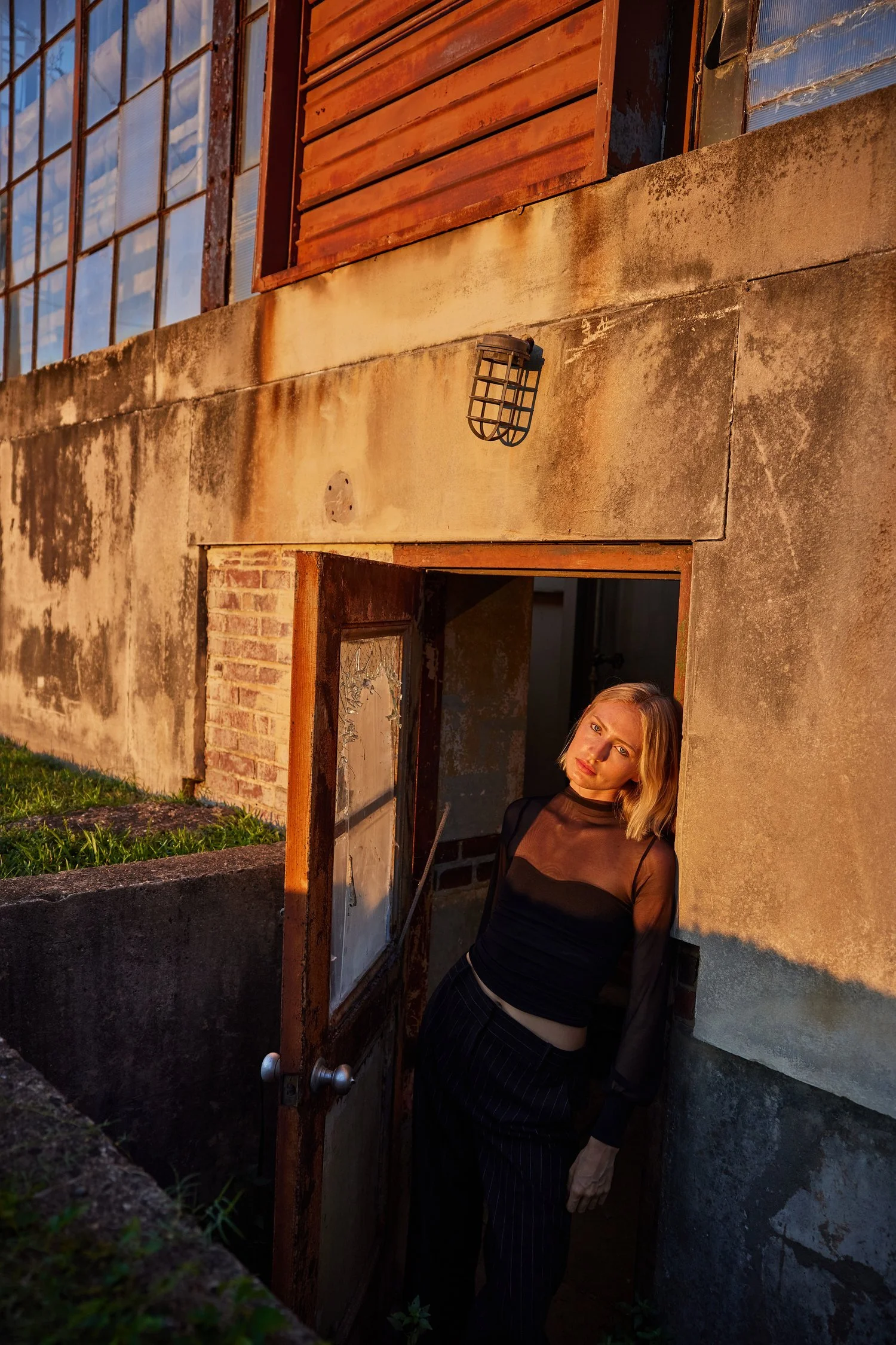 A woman with blonde hair wearing a black top and black pinstripe pants standing in a doorway of an industrial building during sunset, with warm golden light on her face.