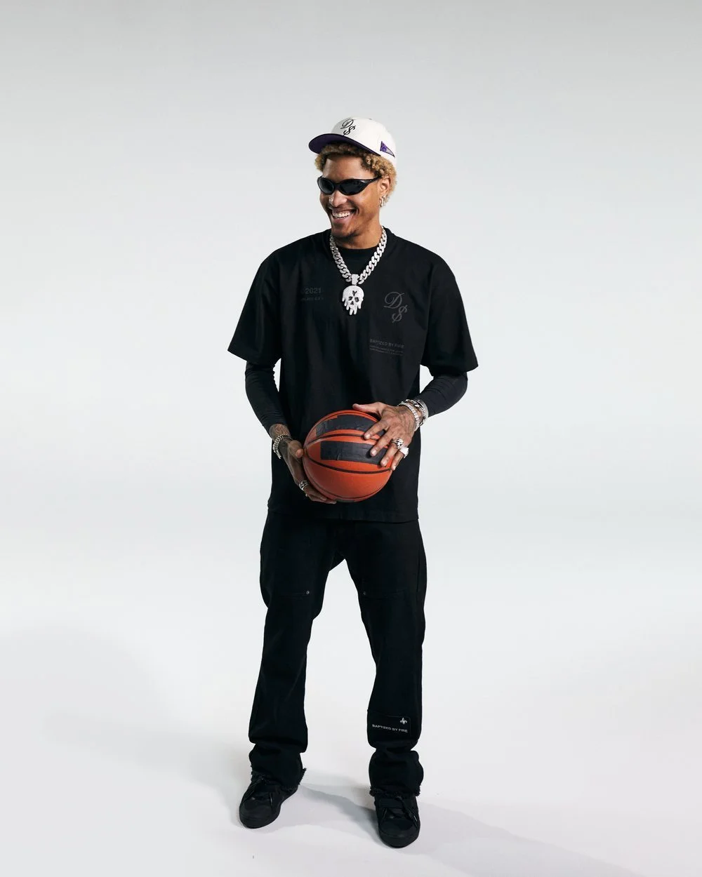 A man dressed in black clothing holding a basketball, wearing sunglasses, a white cap, and jewelry, smiling against a plain background.