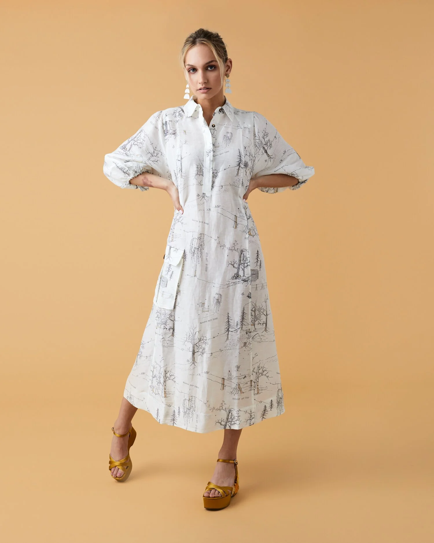 A woman wearing a white long-sleeve dress with a black line drawing print, standing with her hands on her hips, against a beige background, wearing tan platform heels and large earrings.