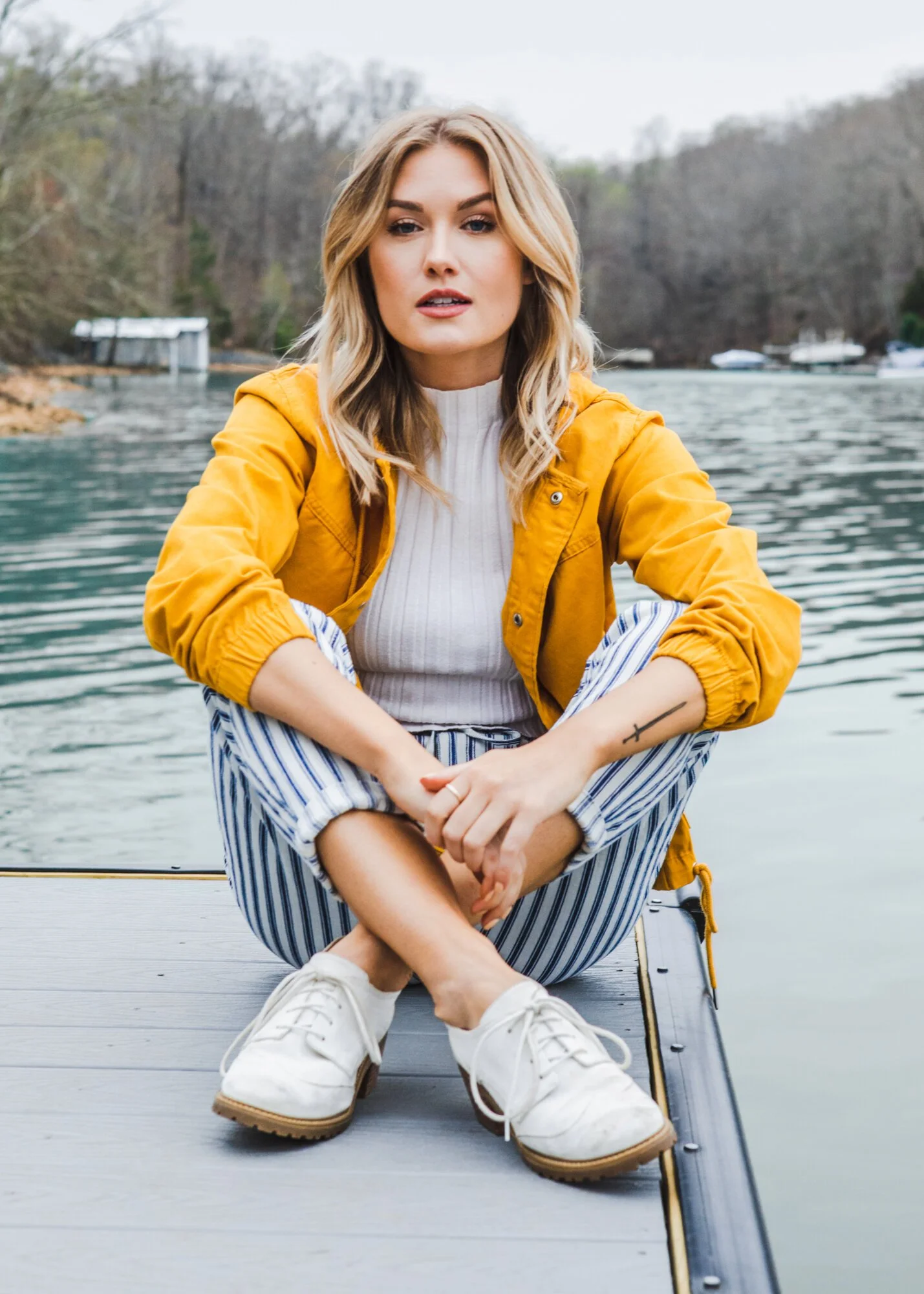 A young woman with blonde hair sits on a dock by the water, wearing a yellow jacket, white shirt, striped pants, and white shoes, with trees and boats in the background.